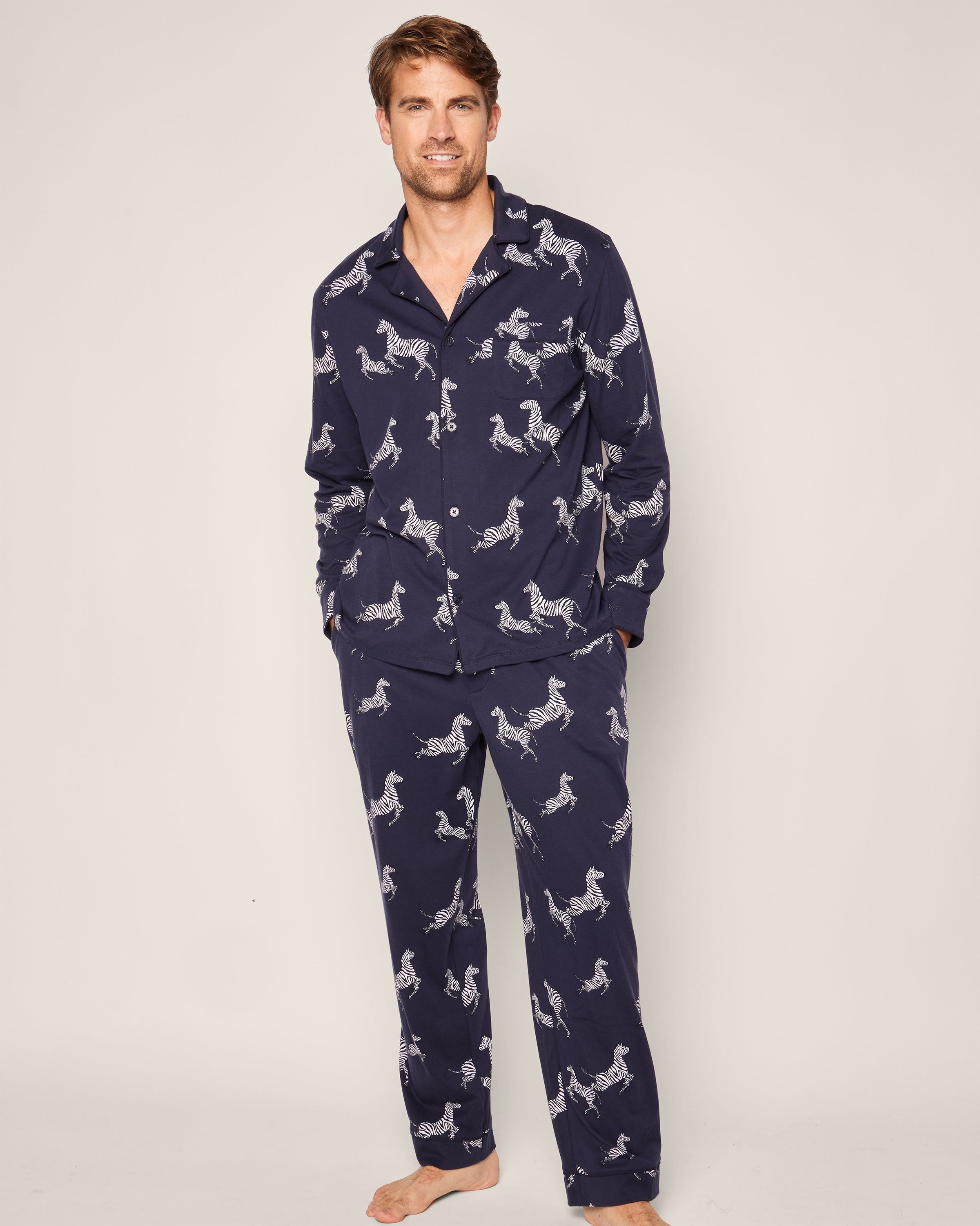 A person stands barefoot in Petite Plumes luxurious Mens Pima Pajama Set in Zèbre de Nuit. The dark blue zebra-print set includes a long-sleeve button-up top and loose pants. They smile warmly, hands casually tucked into pockets.