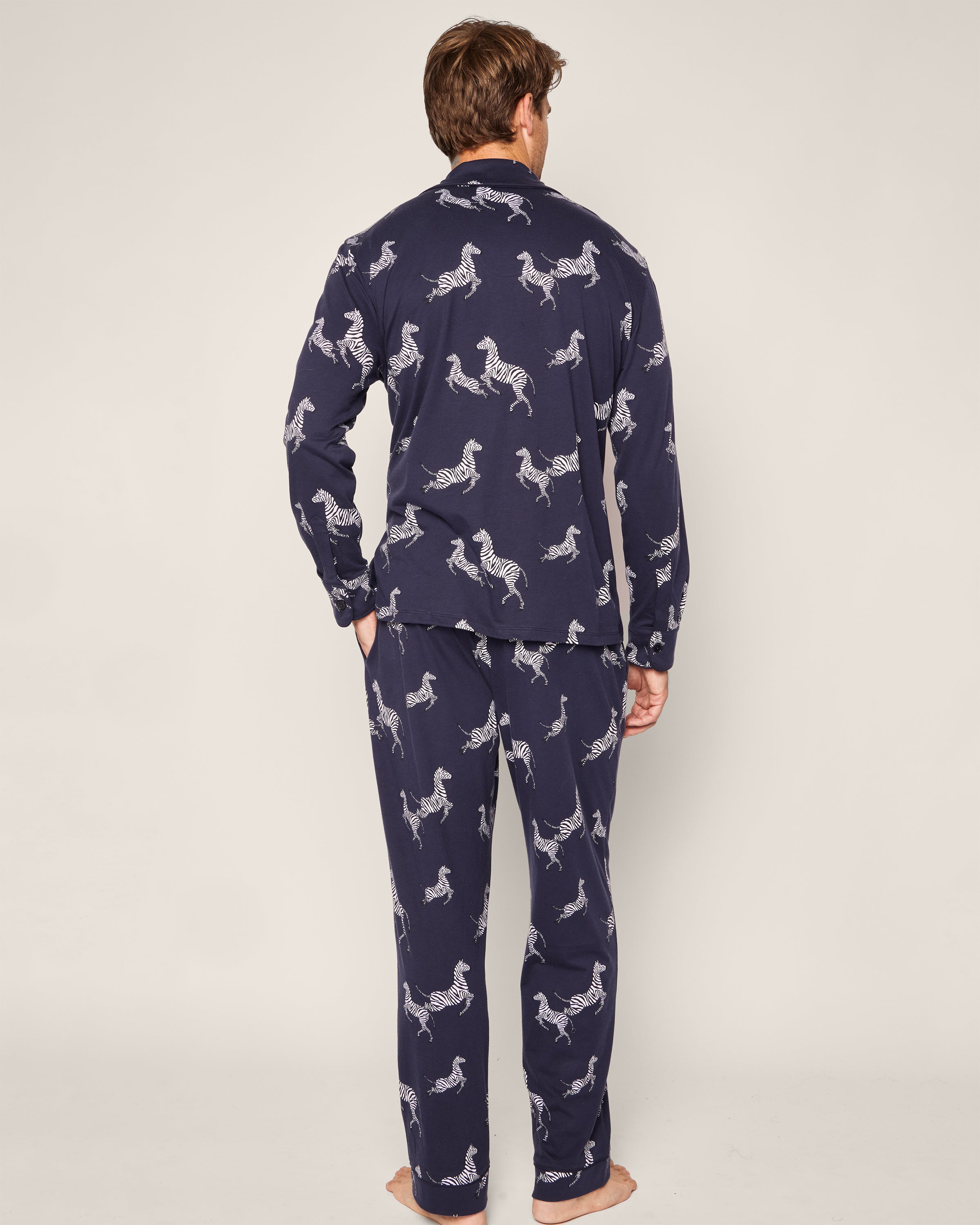 Facing away, a person wears the luxurious Mens Pima Pajama Set in Zèbre de Nuit by Petite Plume—an elegant navy pajama set with a white zebra print. A plain, light background highlights this striking design.