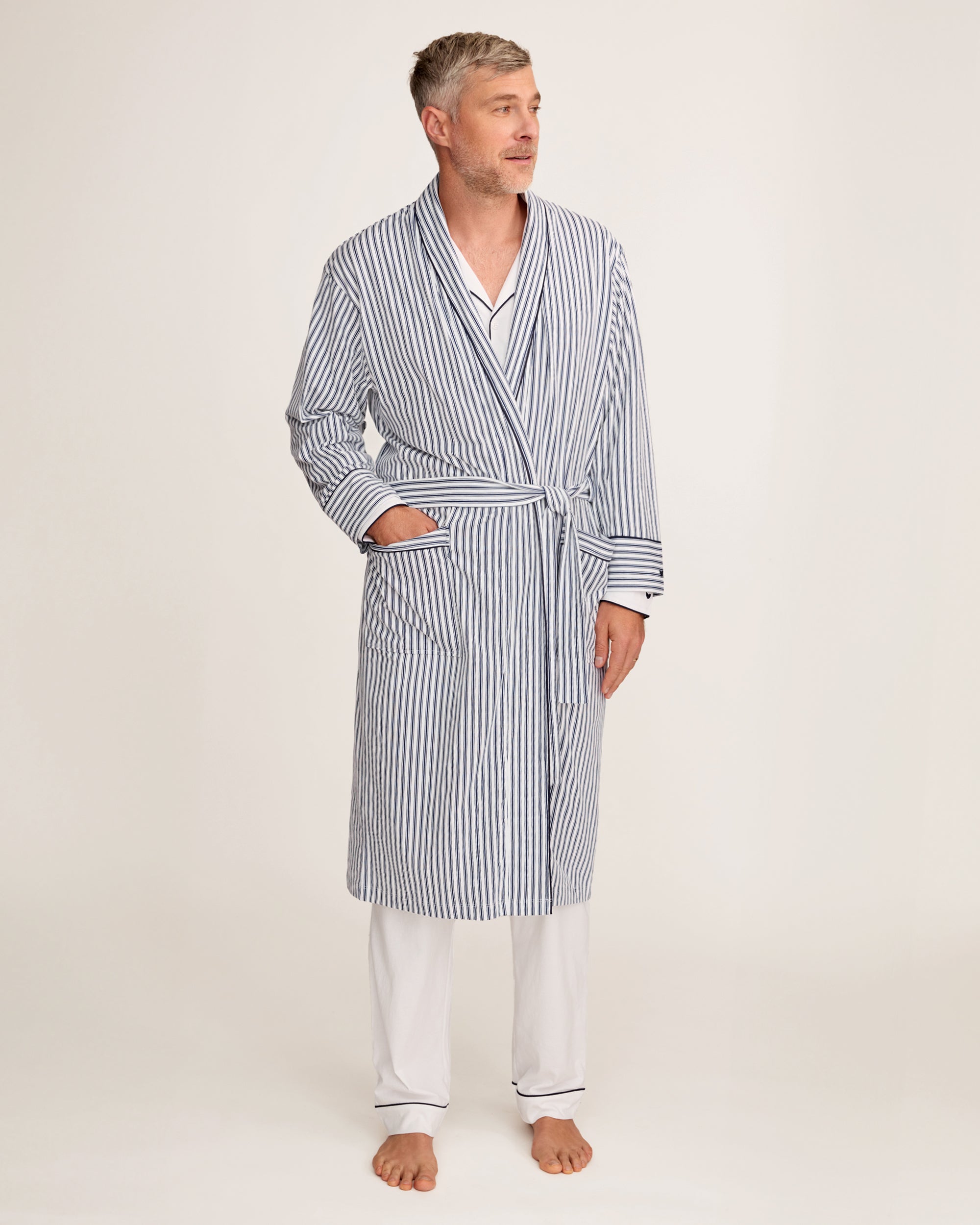 A man stands barefoot in white pajamas and the Men's Pima Robe in Navy French Ticking, belted, looking to the side against a plain light background.