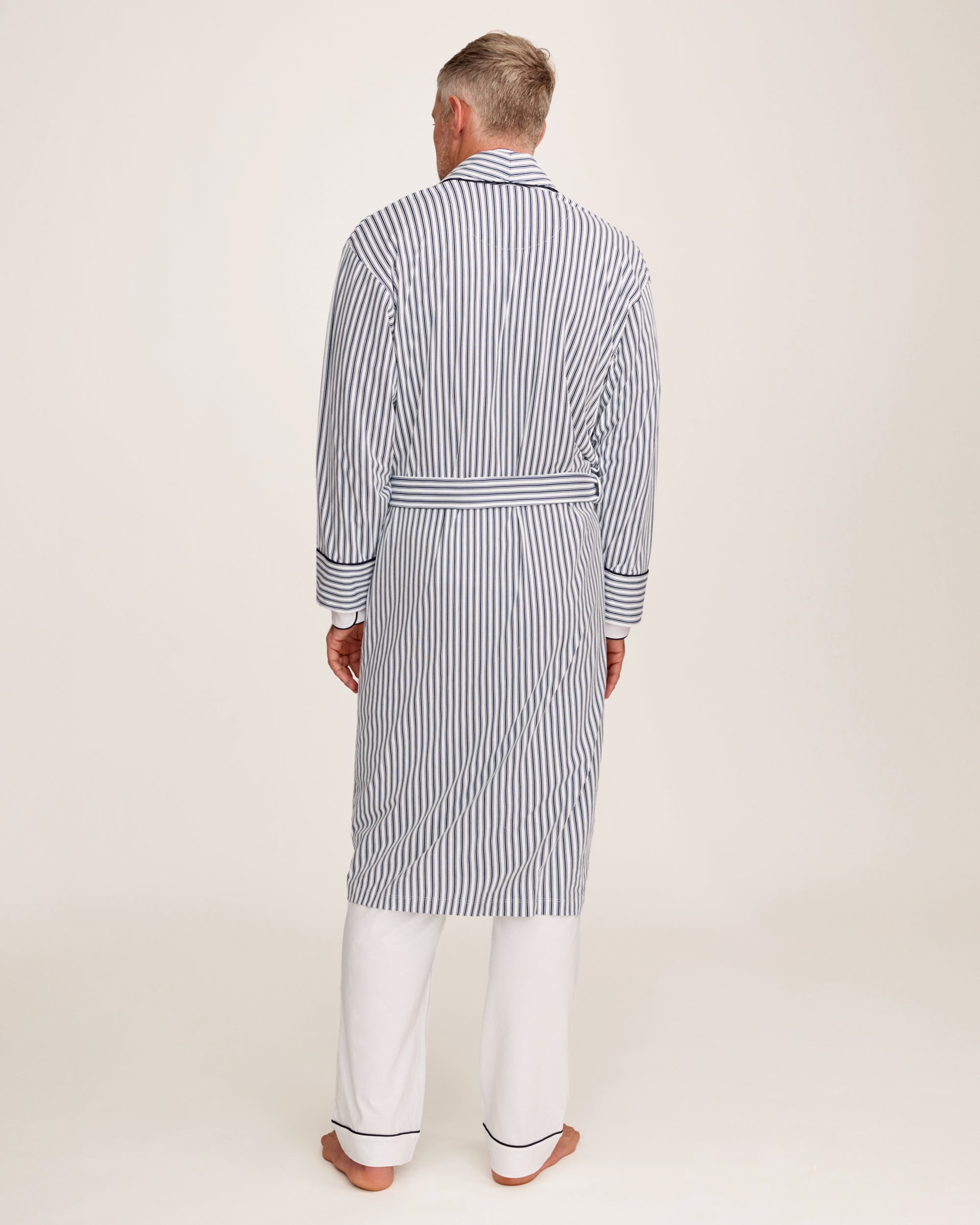 A man stands barefoot in white pajamas and the Men's Pima Robe in Navy French Ticking, belted, looking to the side against a plain light background.