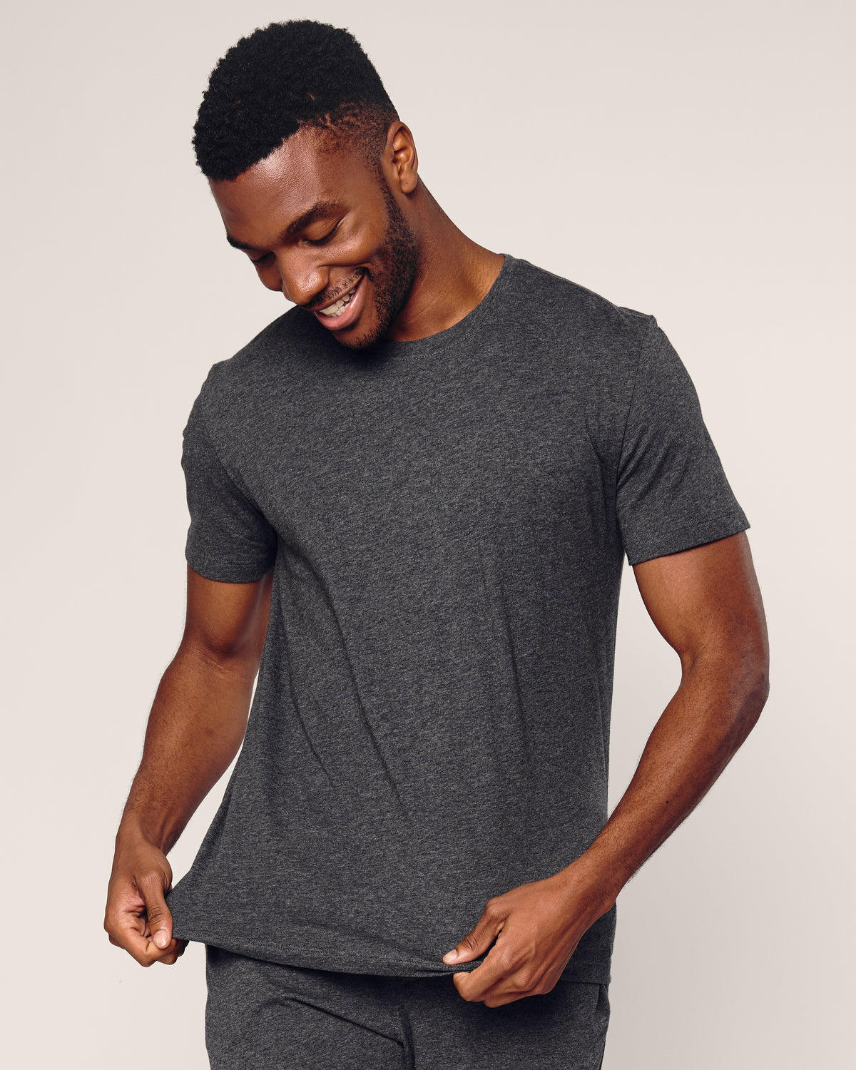 A man in a Petite Plume Mens Pima Crew Neck Short Sleeve Top in Dark Grey smiles and gently tugs the hem of his shirt against a light neutral background.