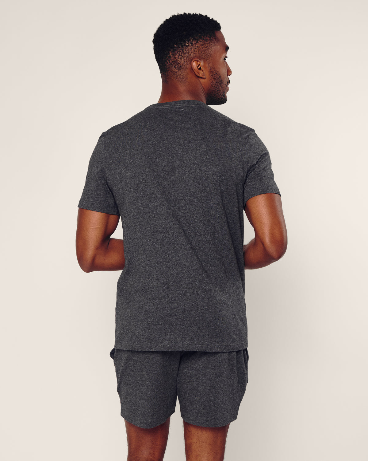 A person faces away from the camera, wearing Petite Plumes Mens Pima Crew Neck Short Sleeve Top in Dark Grey and matching shorts against an off-white background. With short black hair, they glance sideways, radiating comfort and style.