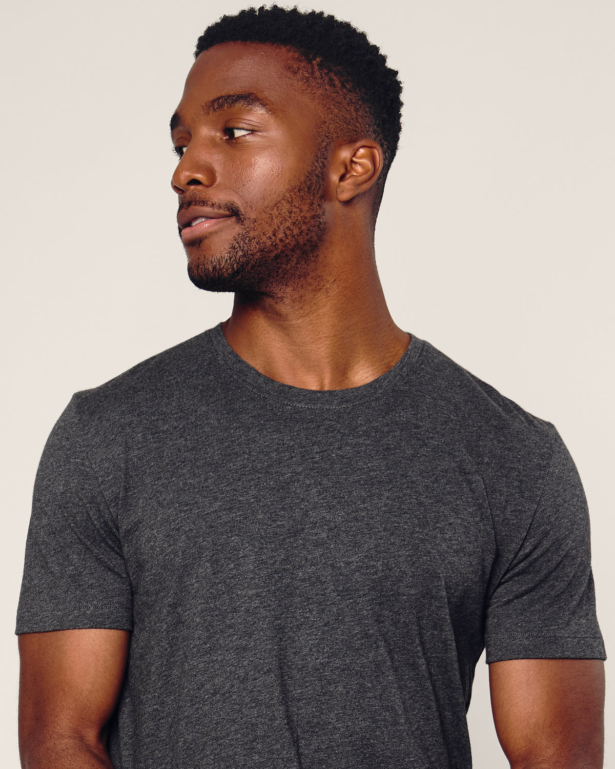 A man with short black hair and a beard wears the Petite Plume Mens Pima Crew Neck Short Sleeve Top in Dark Grey, contemplating his bedtime ritual while looking to his left against a plain, light-colored background.