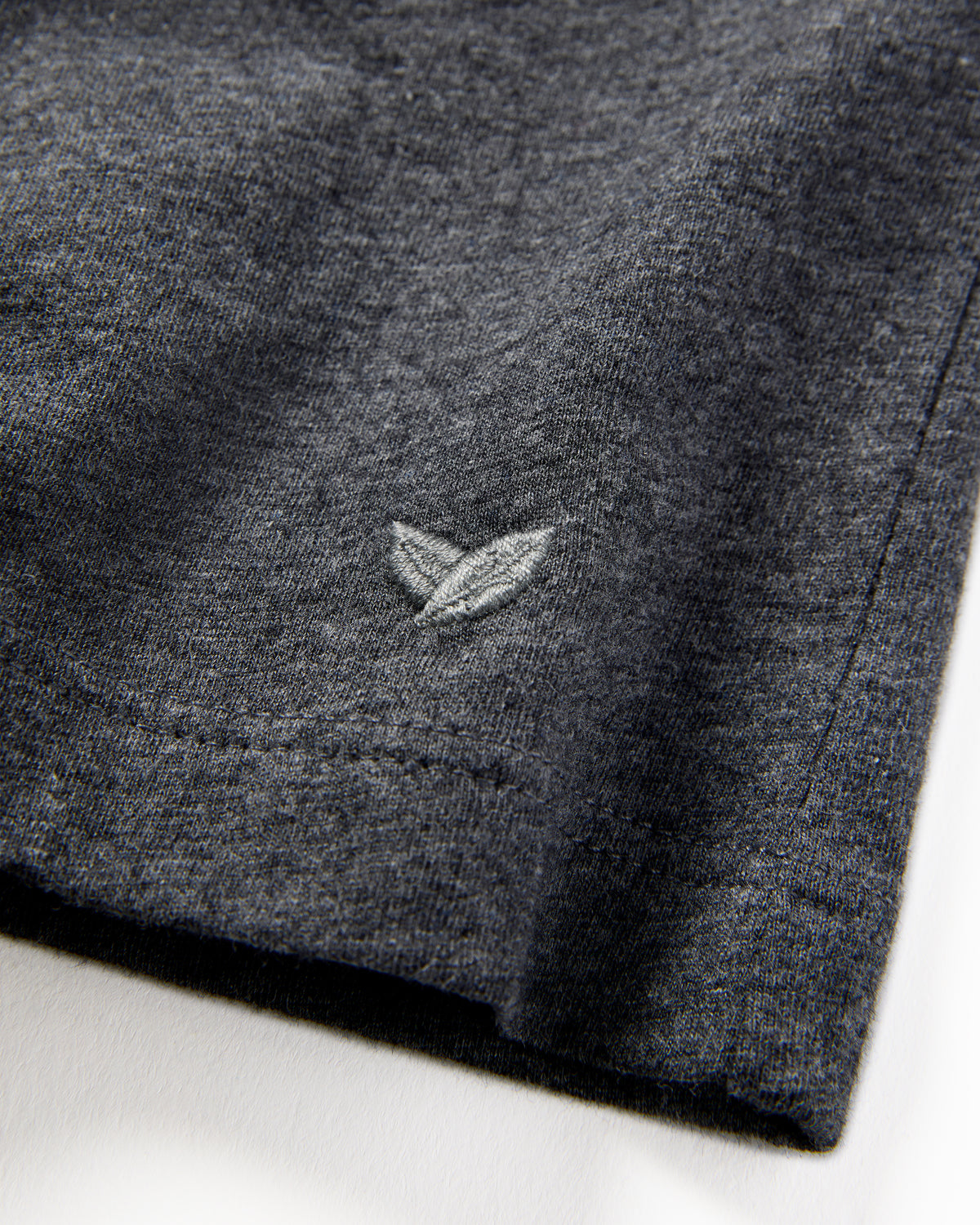 Close-up of Petite Plumes Mens Pima Crew Neck Short Sleeve Top in Dark Grey, crafted from Peruvian Pima cotton, highlighting a small leaf-like embroidered logo. The luxurious texture and stitching details around the logo are visible.