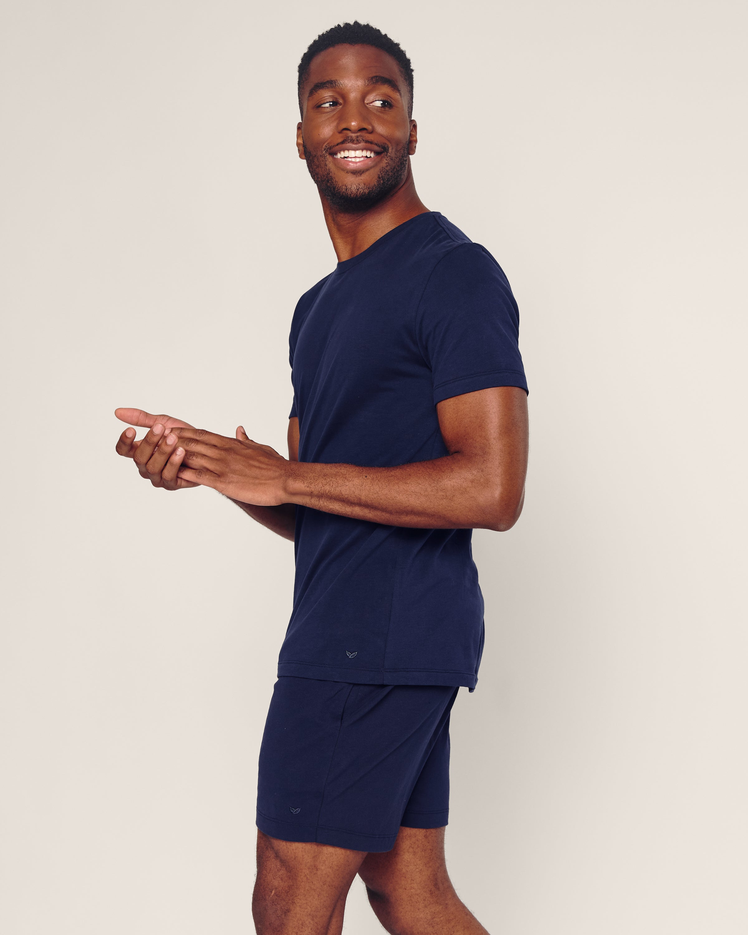 A man wearing Petite Plumes Mens Pima Crew Neck Short Sleeve Top in Navy and matching shorts smiles and glances to the side, standing against a plain, light-colored background.