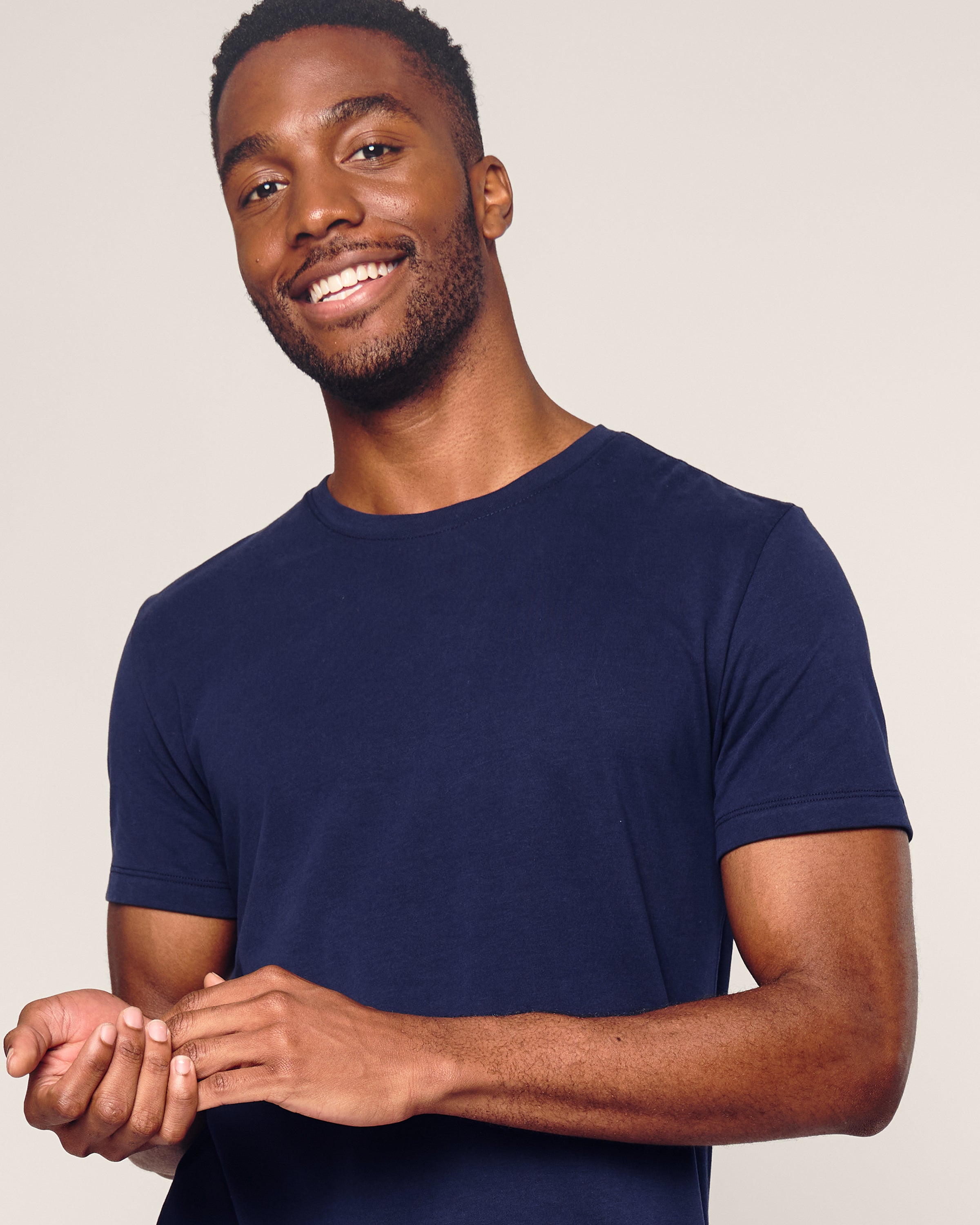 A smiling man in a Petite Plume Mens Pima Crew Neck Short Sleeve Top in Navy stands against a plain light background. The soft fabric suggests the comfort of jersey knit pajamas. His short hair and beard complete the relaxed look as he holds something gently in his hands.