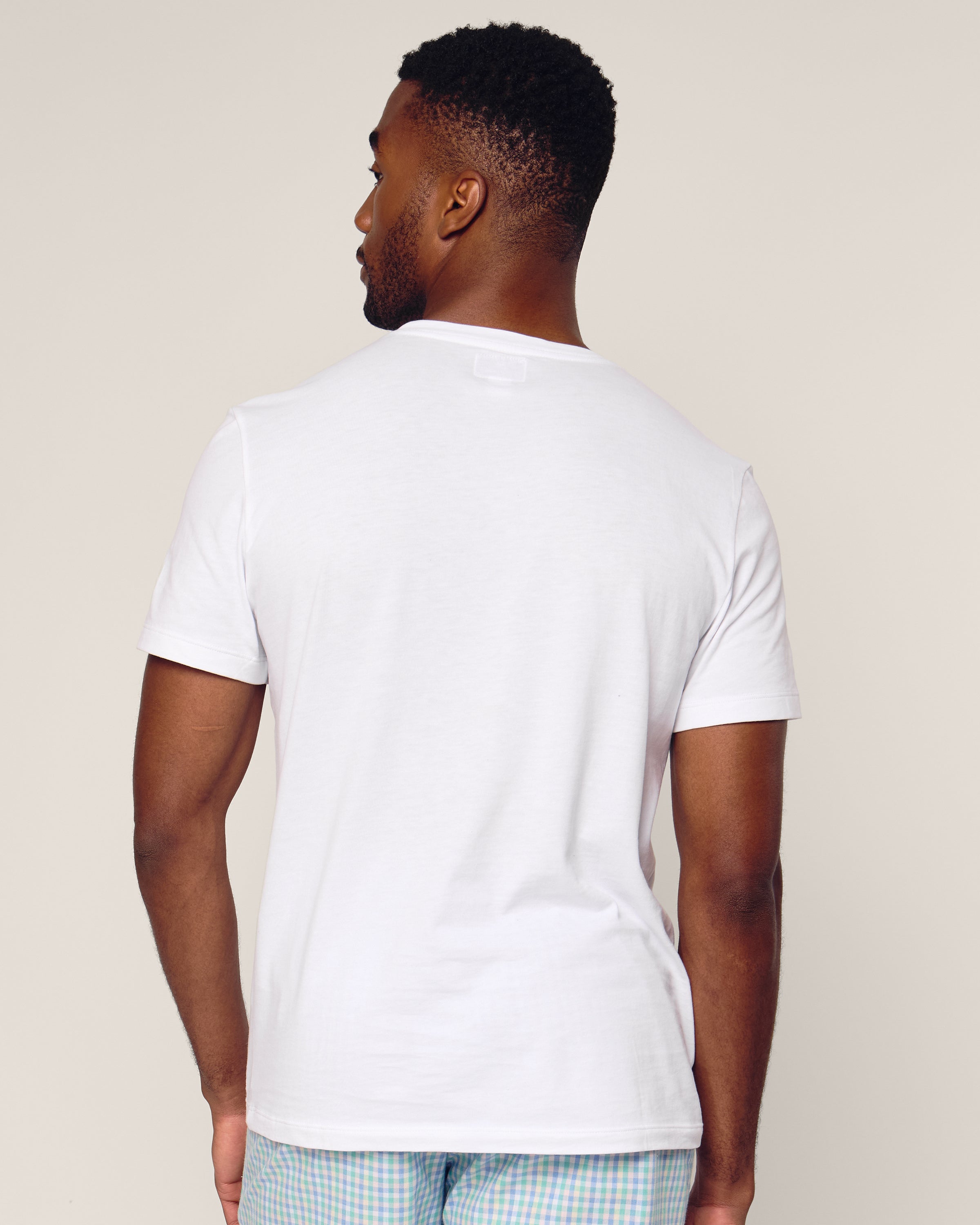 A person stands with their back to the camera, wearing Petite Plumes Mens Pima Crew Neck Short Sleeve Top in White and light-colored plaid pants made of soft Peruvian Pima cotton against an off-white background.