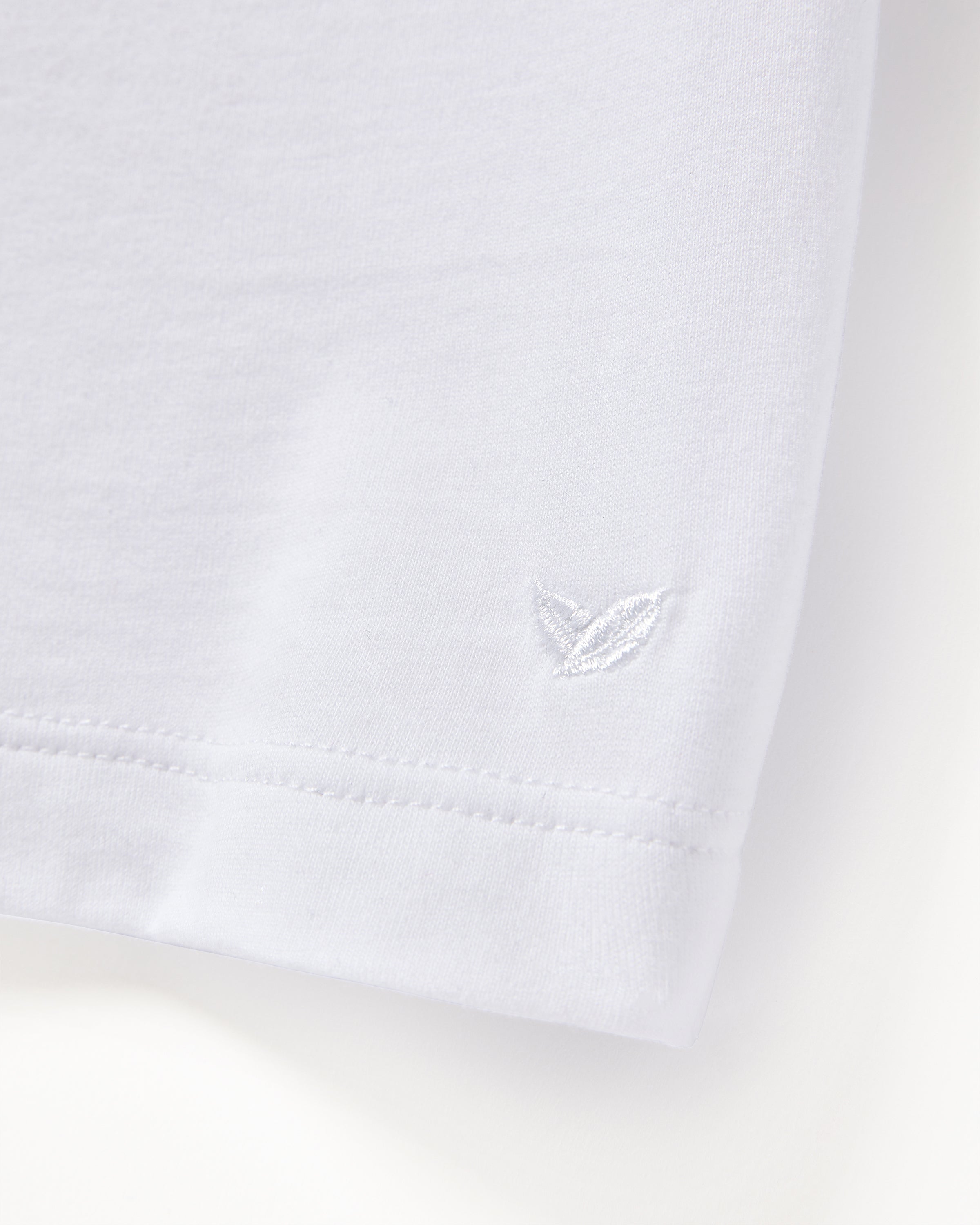 A close-up of the Mens Pima Crew Neck Short Sleeve Top in White by Petite Plume reveals luxurious Peruvian Pima cotton fabric with delicate stitching near the hem and a small white embroidered leaf on the smooth jersey knit.