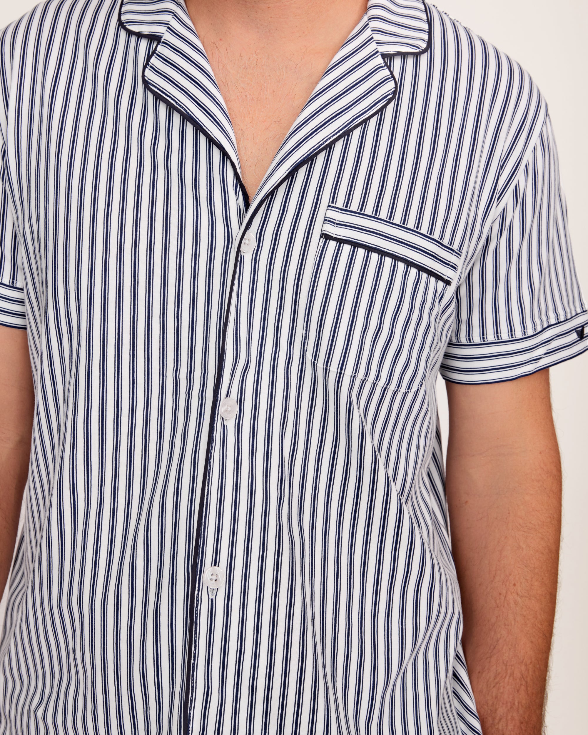 A man stands barefoot in a studio wearing the Men's Pima Pajama Short Set in Navy French Ticking, which features vertical blue and white stripes. He looks slightly to the side with one hand in his pocket.