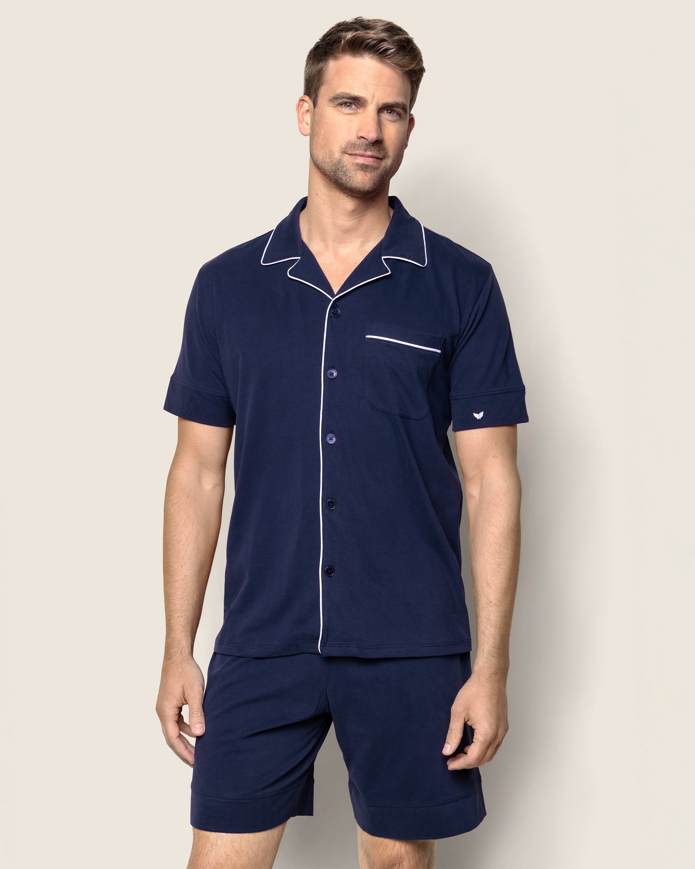 A man is wearing the Mens Pima Pajama Short Set in Navy by Petite Plume, a luxurious Peruvian Pima cotton short-sleeve set with white piping, button-up top, and pocket. He stands against a plain light background.