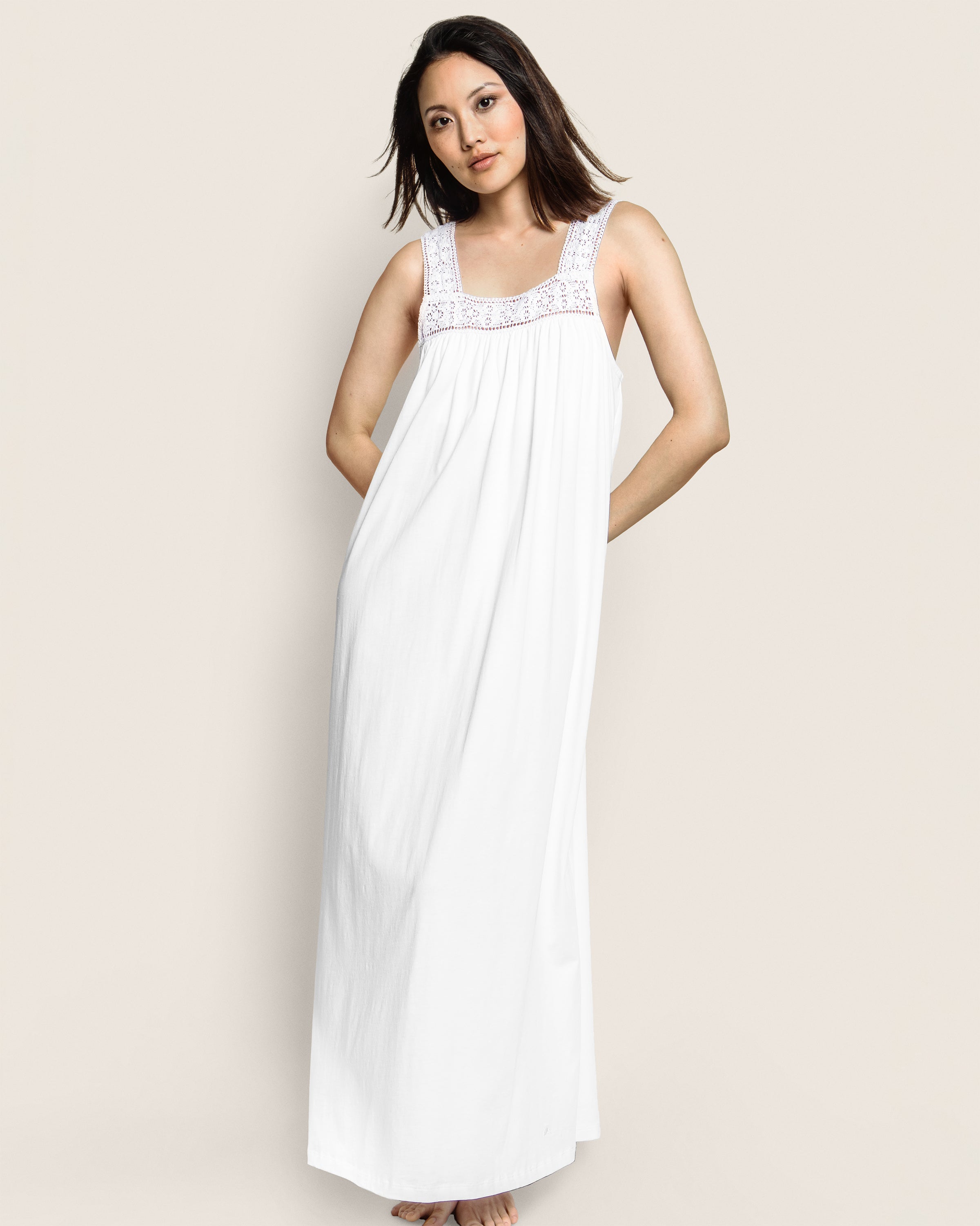 A woman poses against a plain background wearing the Womens Pima Camille Nightgown in White by Petite Plume, featuring a decorative lace neckline. Her hands rest behind her back, and her hair is down, softly blowing to one side.