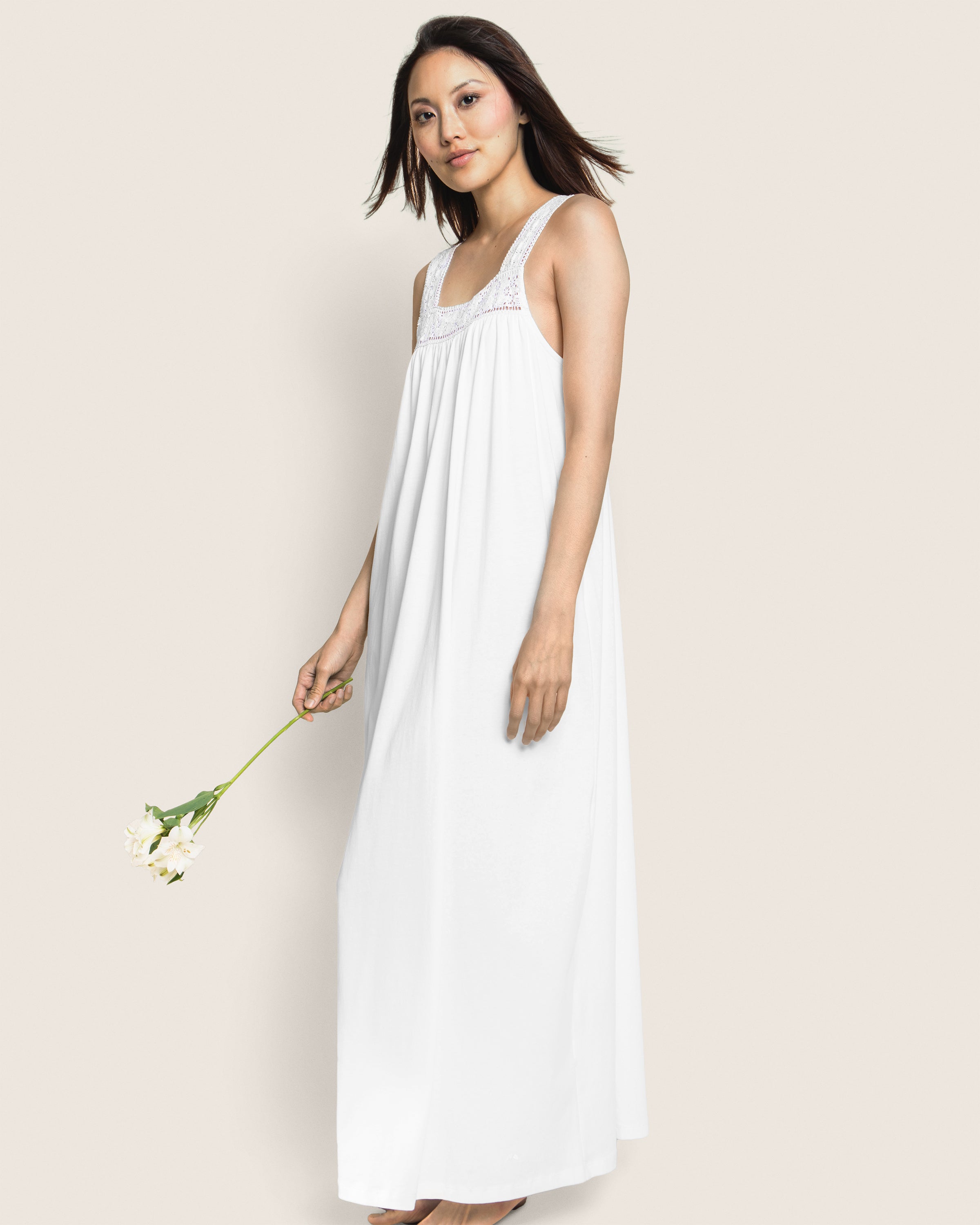 A woman in a flowing white nightgown, embodying the elegance of Petite Plumes Womens Pima Camille collection, stands with a single white flower. Her hair gently blows to one side against a plain backdrop, highlighting the simple sophistication of Peruvian Pima cotton.
