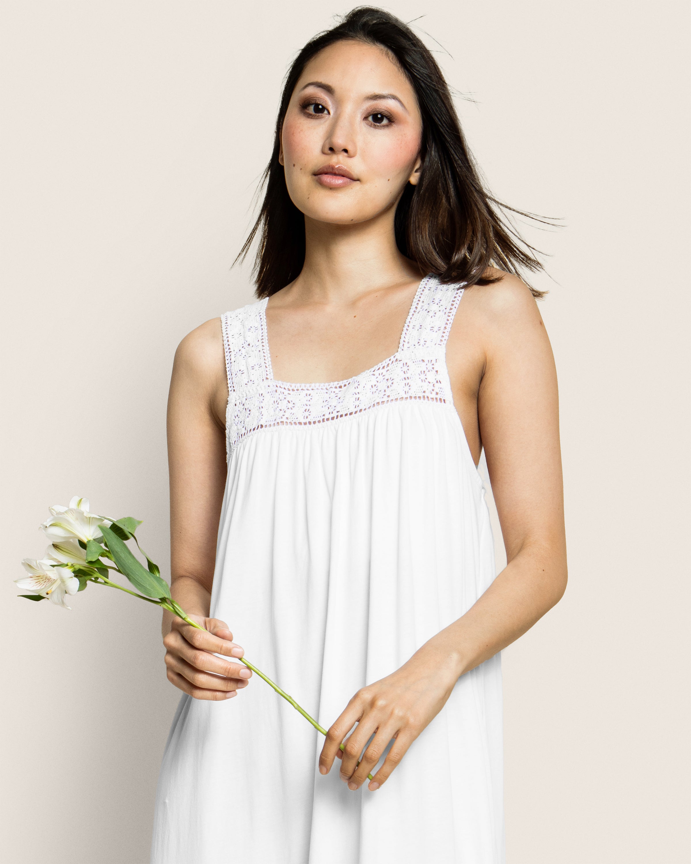 A woman in a white, sleeveless dress with lace details holds a bouquet of white flowers, akin to the elegance of Petite Plumes Womens Pima Camille Nightgown in White. She has shoulder-length hair and gazes calmly at the camera against a plain background.