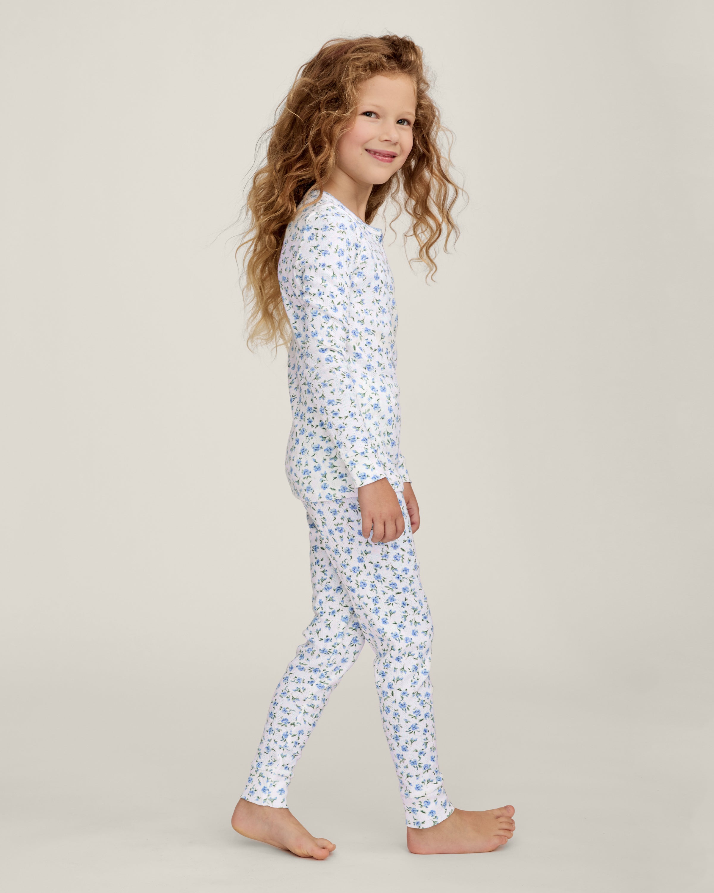 A young child with long curly hair stands barefoot against a plain background, wearing the Kid's Pointelle Snug Fit Pajama Set in Blue Floral.