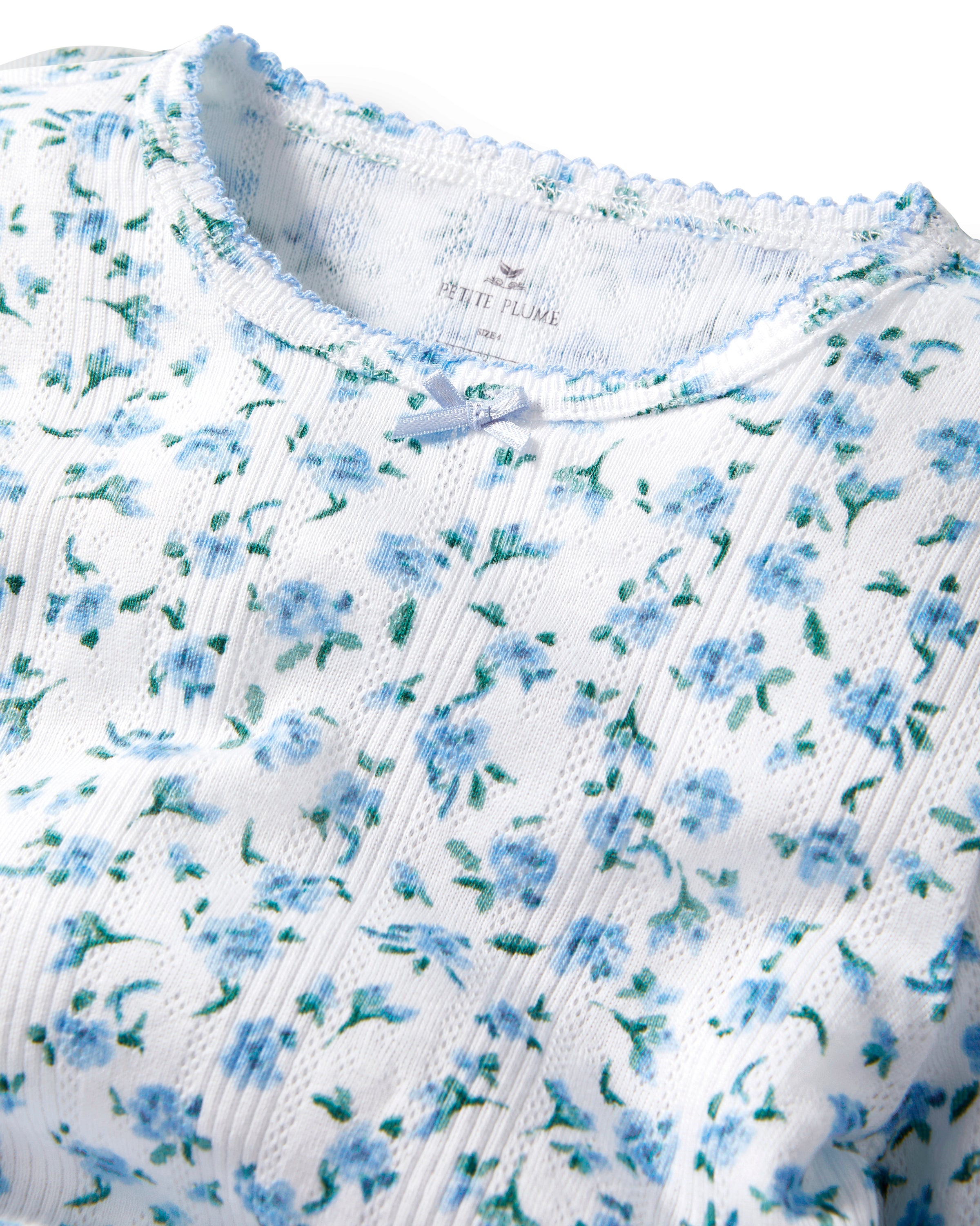 A young child with long curly hair stands barefoot against a plain background, wearing the Kid's Pointelle Snug Fit Pajama Set in Blue Floral.