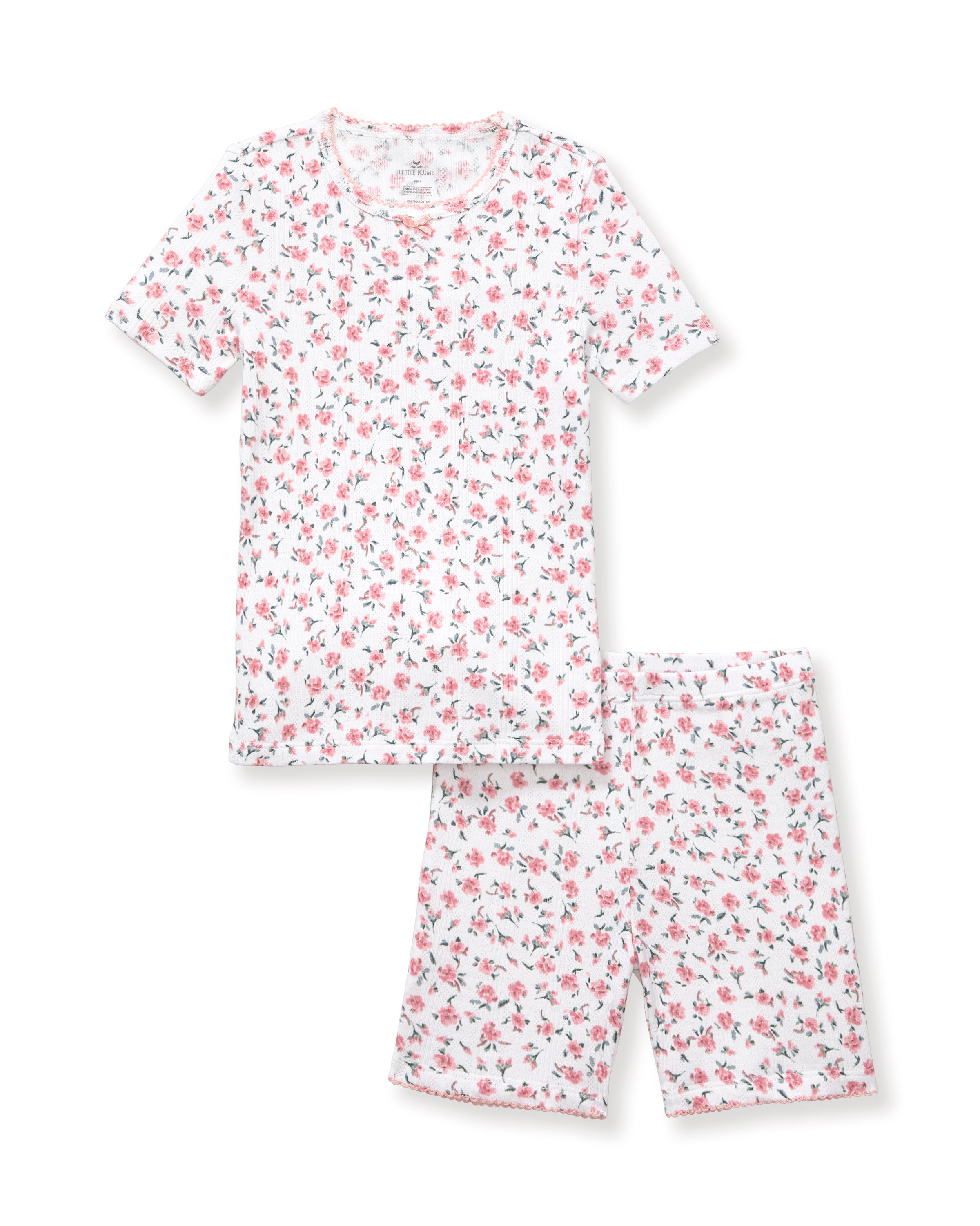A young girl with curly hair stands against a plain background, wearing the Kid's Pointelle Snug Fit Pajama Short Set in Pink Floral, featuring a matching short-sleeve top and shorts decorated with a pink and red floral pattern.