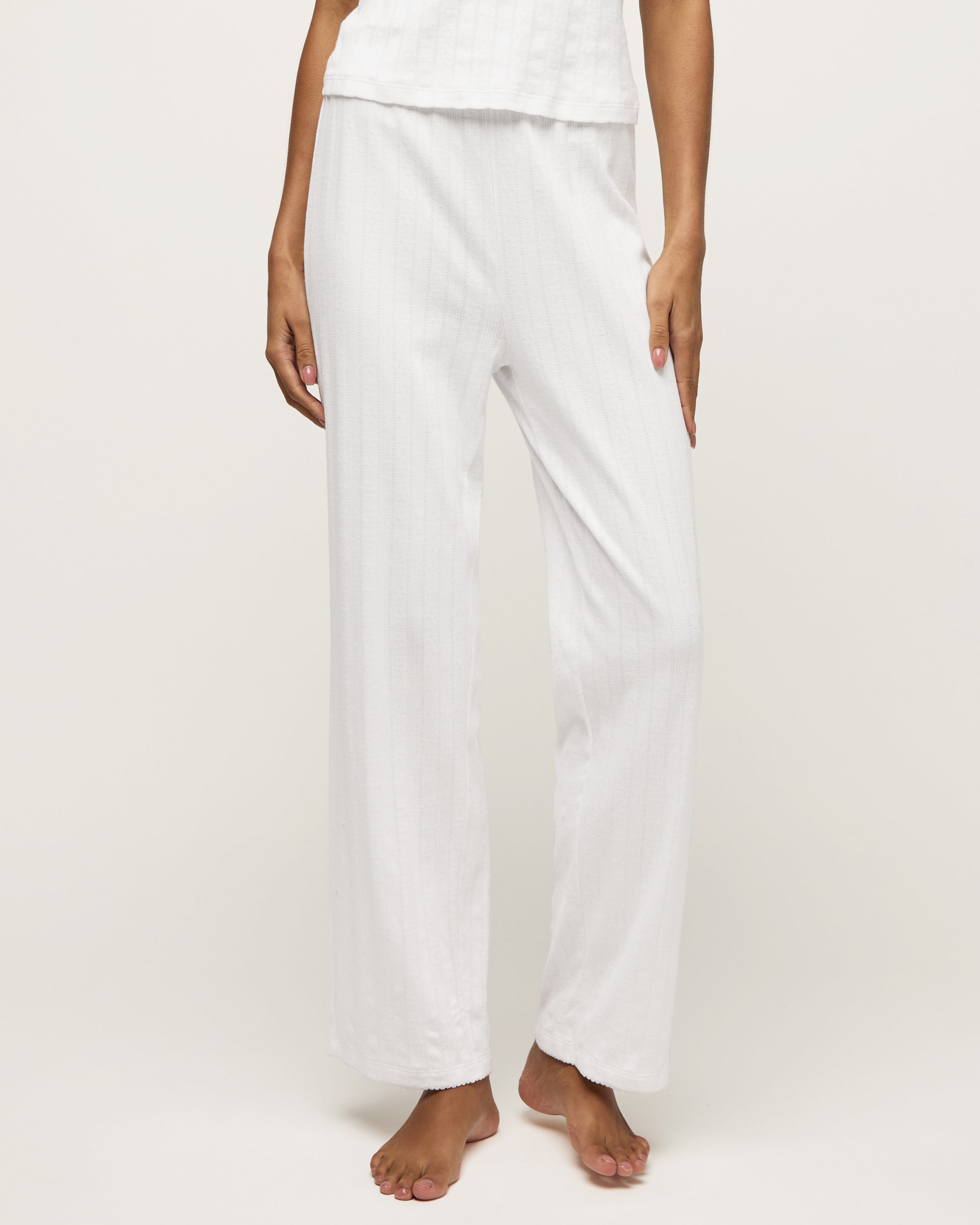 A woman stands barefoot on a light background wearing the Women's Pointelle Pant in White, paired with a sleeveless white tank top, posing with a relaxed and neutral expression.