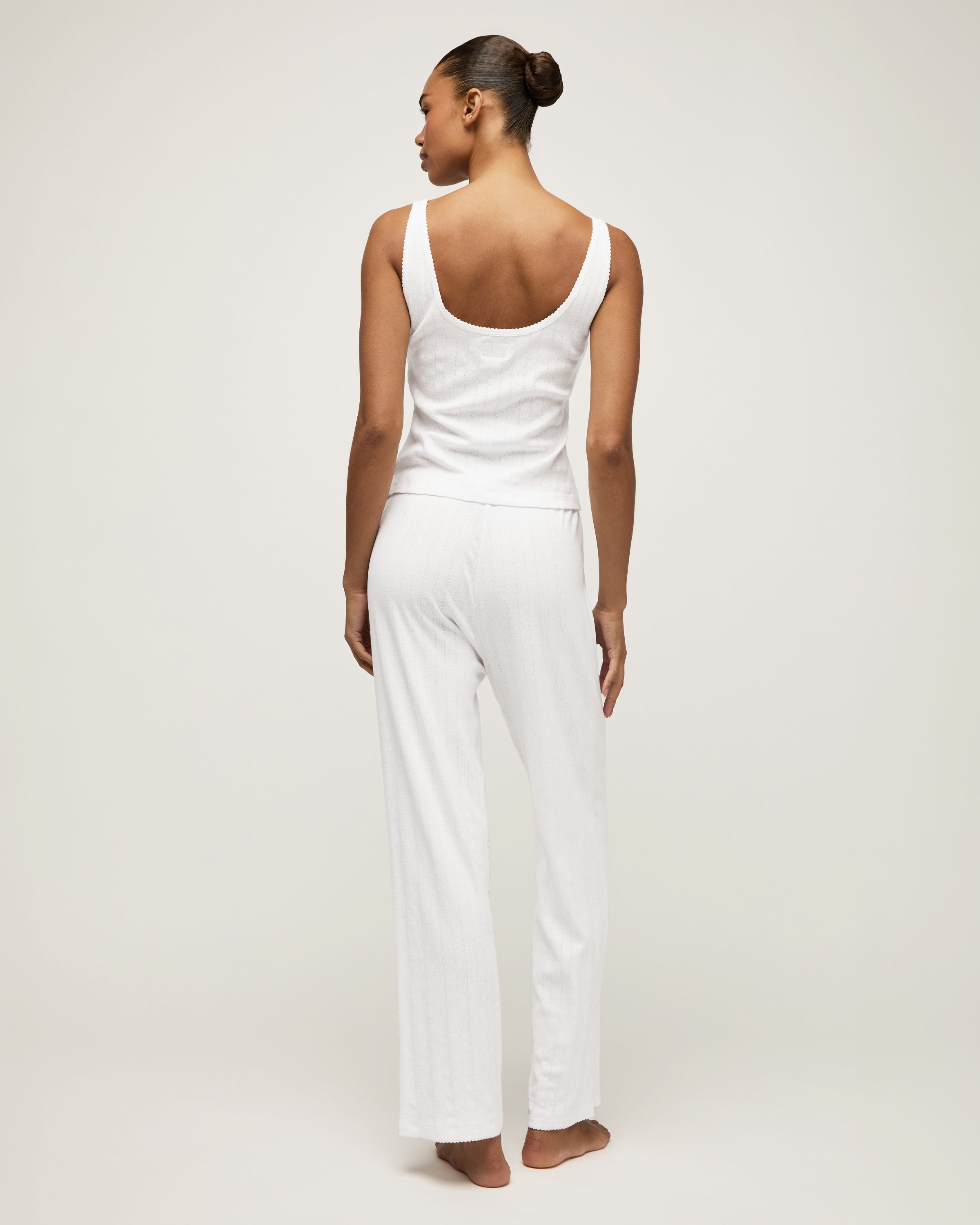 A woman stands barefoot on a light background wearing the Women's Pointelle Pant in White, paired with a sleeveless white tank top, posing with a relaxed and neutral expression.