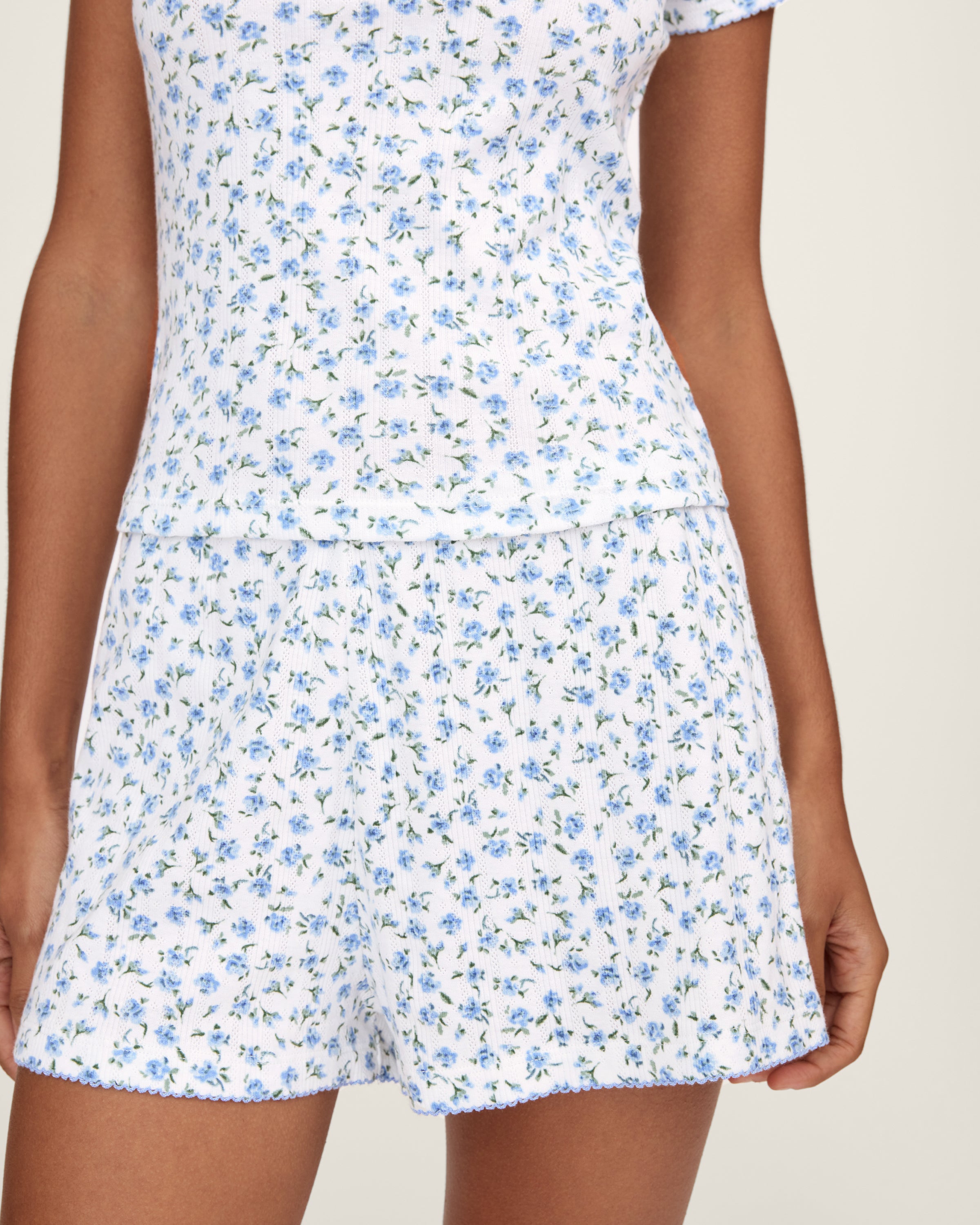 A person stands barefoot against a plain background, wearing Women's Pointelle Shorts in Blue Floral with a matching short-sleeve top. They have long, wavy hair and a relaxed, neutral expression.