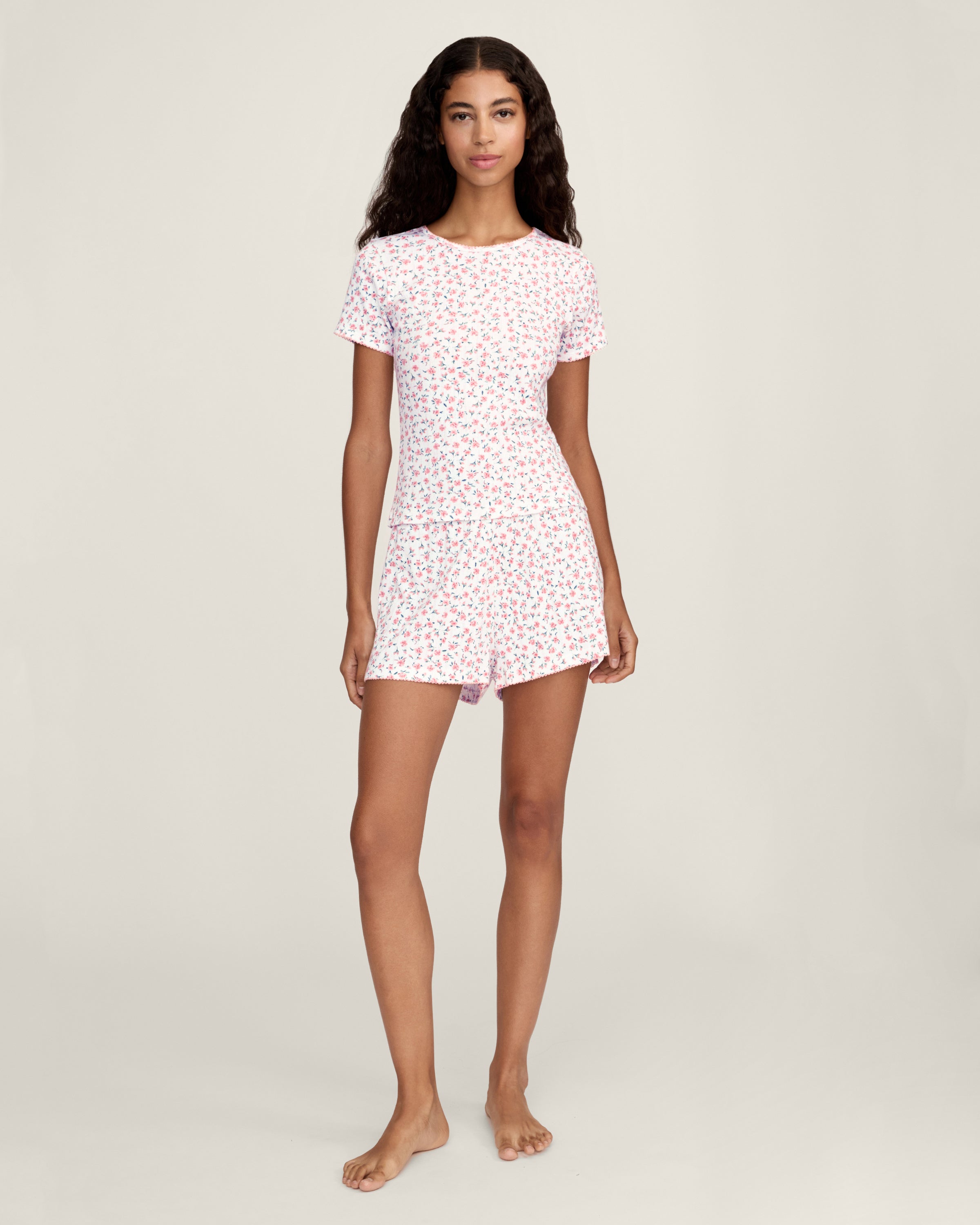 A woman with long curly hair and a relaxed expression stands barefoot against a neutral background, wearing Women's Pointelle Shorts in Pink Floral as part of a matching short-sleeved pajama set.