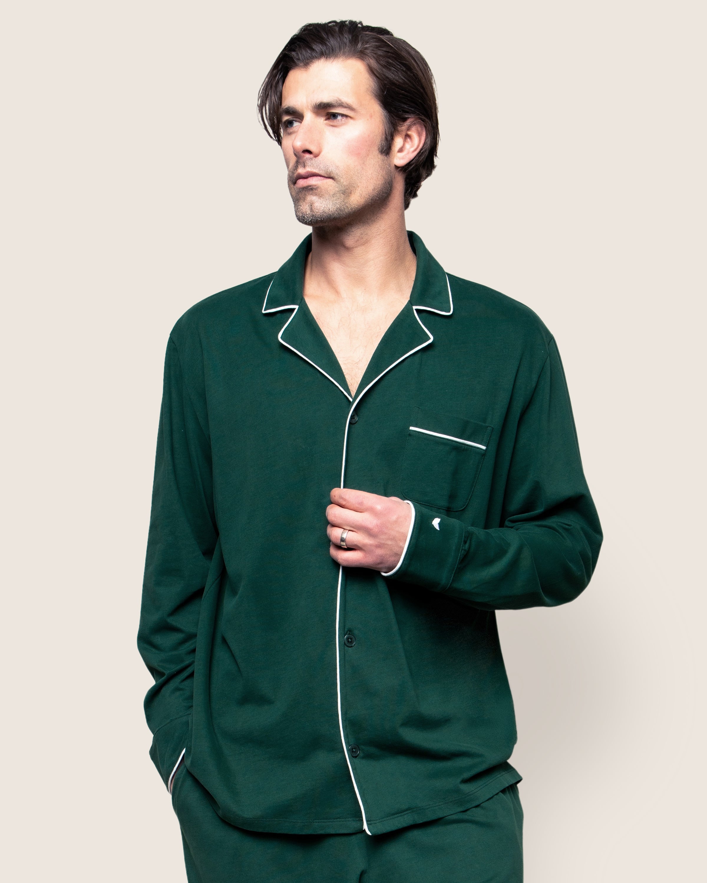 The Mens Pima Pajama Set in Evergreen by Petite Plume is crafted from luxury Peruvian Pima cotton. This timeless set includes a dark green long-sleeve button-up top with white piping and a chest pocket, paired with matching pants featuring an elastic waistband, on a pristine white background.