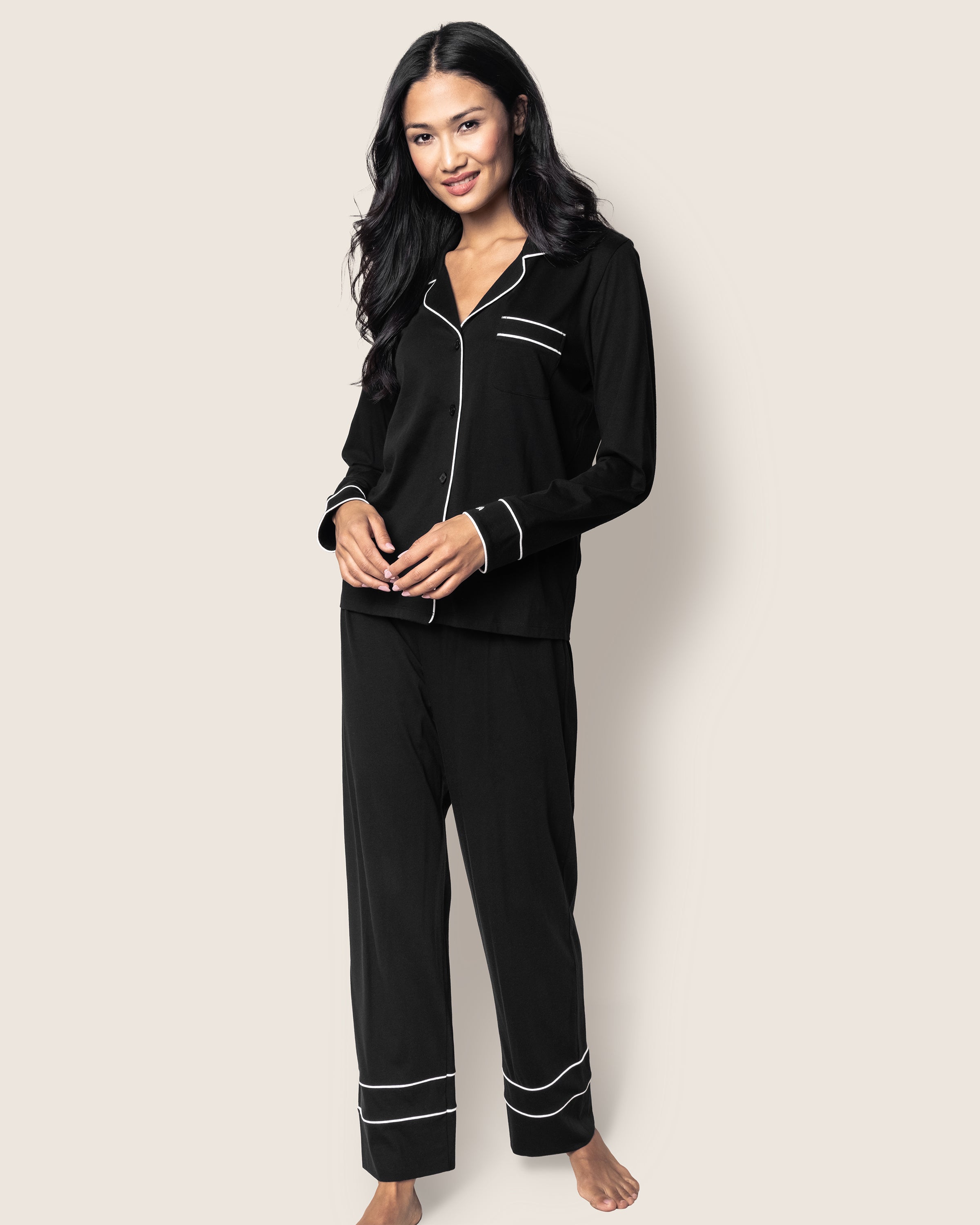 A long-haired person stands barefoot against a plain background, wearing the Womens Pima Astaire Pajama Set in Black by Petite Plume. The luxury pajamas are yarn-dyed with white piping on the cuffs, pockets, and edges. They smile with hands clasped in front, radiating comfort and style.