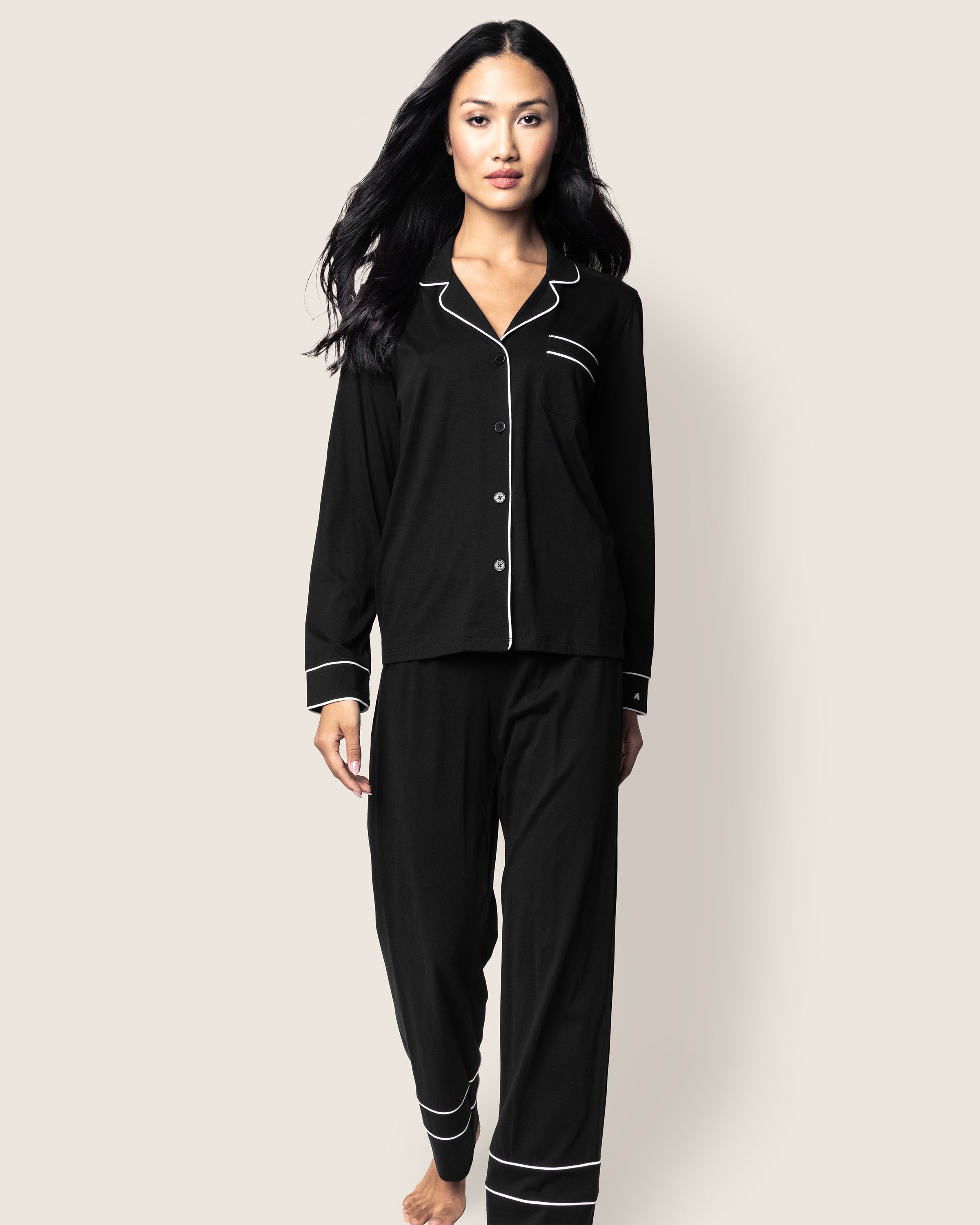 A person with long dark hair walks forward in a luxurious Womens Pima Astaire Pajama Set in Black, featuring Peruvian Pima cotton and white piping, by Petite Plume. The background is plain and light-colored.