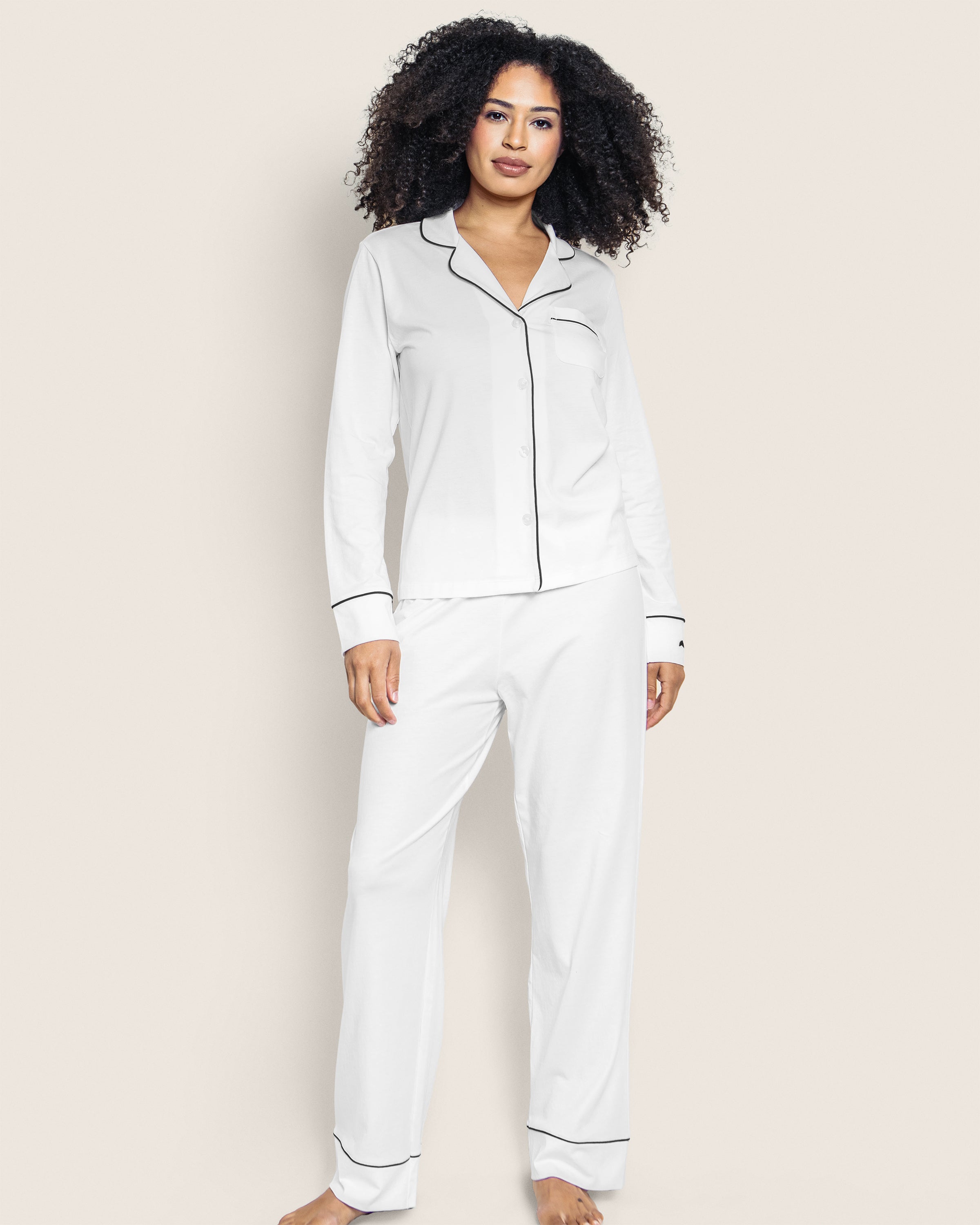 A curly-haired person is wearing Petite Plumes Womens Pima Pajama Set in White with Black Piping, standing barefoot against a plain light background, exuding comfort and style.