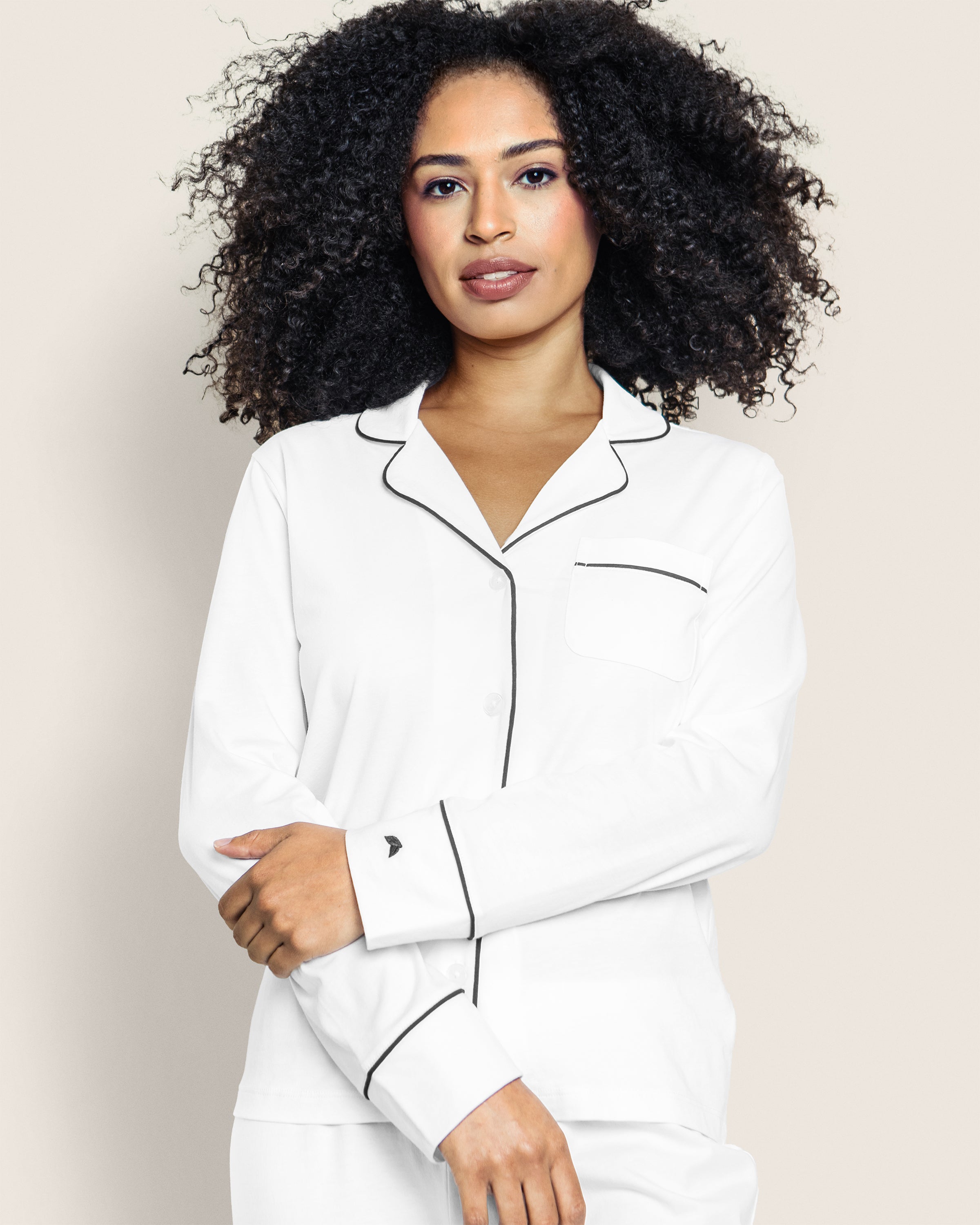 A person with curly hair models the Petite Plume Womens Pima Pajama Set in White with Black Piping. Against a light background, they cross their arms slightly, showcasing the luxurious Peruvian Pima cotton and left-side pocket, embodying relaxed elegance in this luxury sleepwear.