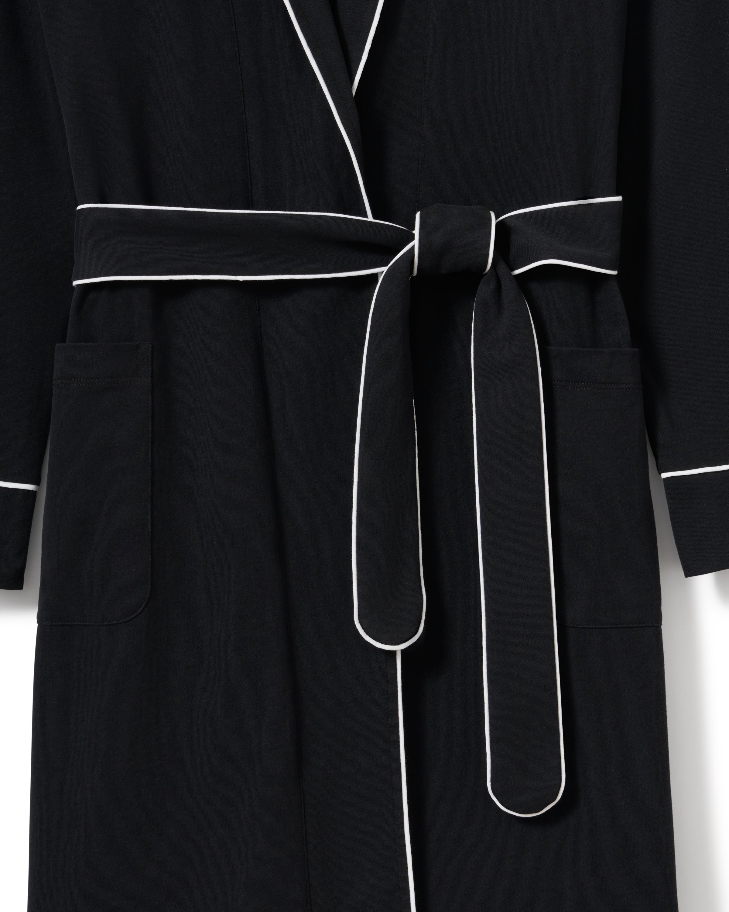A woman wears a Petite Plume Womens Pima Robe in Black, characterized by white piping, long sleeves, and a waist tie. Made from soft Peruvian Pima cotton, she rests her hands in the front pockets while standing against a plain light gray background.
