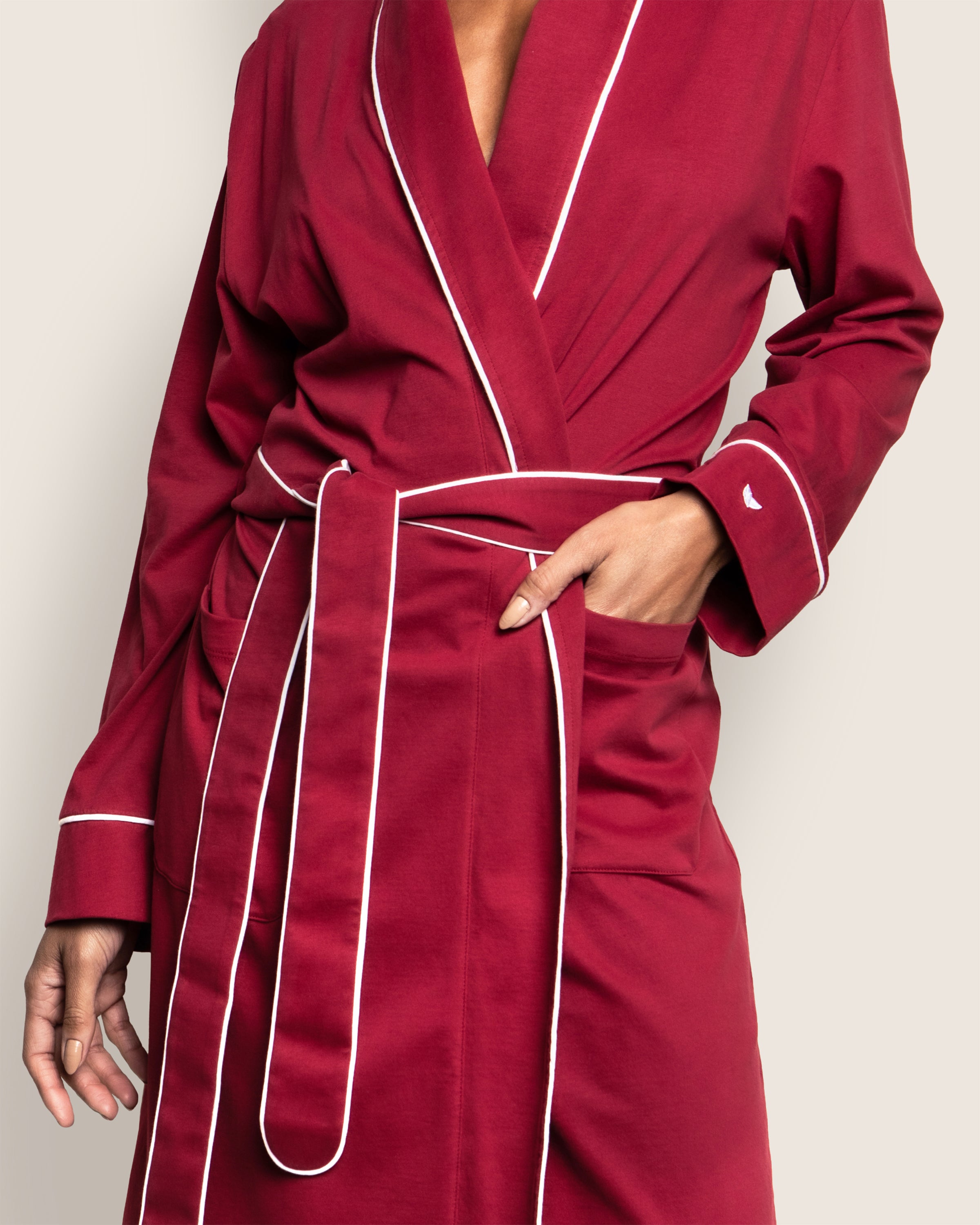 A person in a luxurious Petite Plume Womens Pima Robe in Bordeaux, made from rich Peruvian Pima cotton with classic pajama silhouettes and white piping, stands elegantly against a neutral background. The robe is belted at the waist, with one hand pocketed and the other at their side.