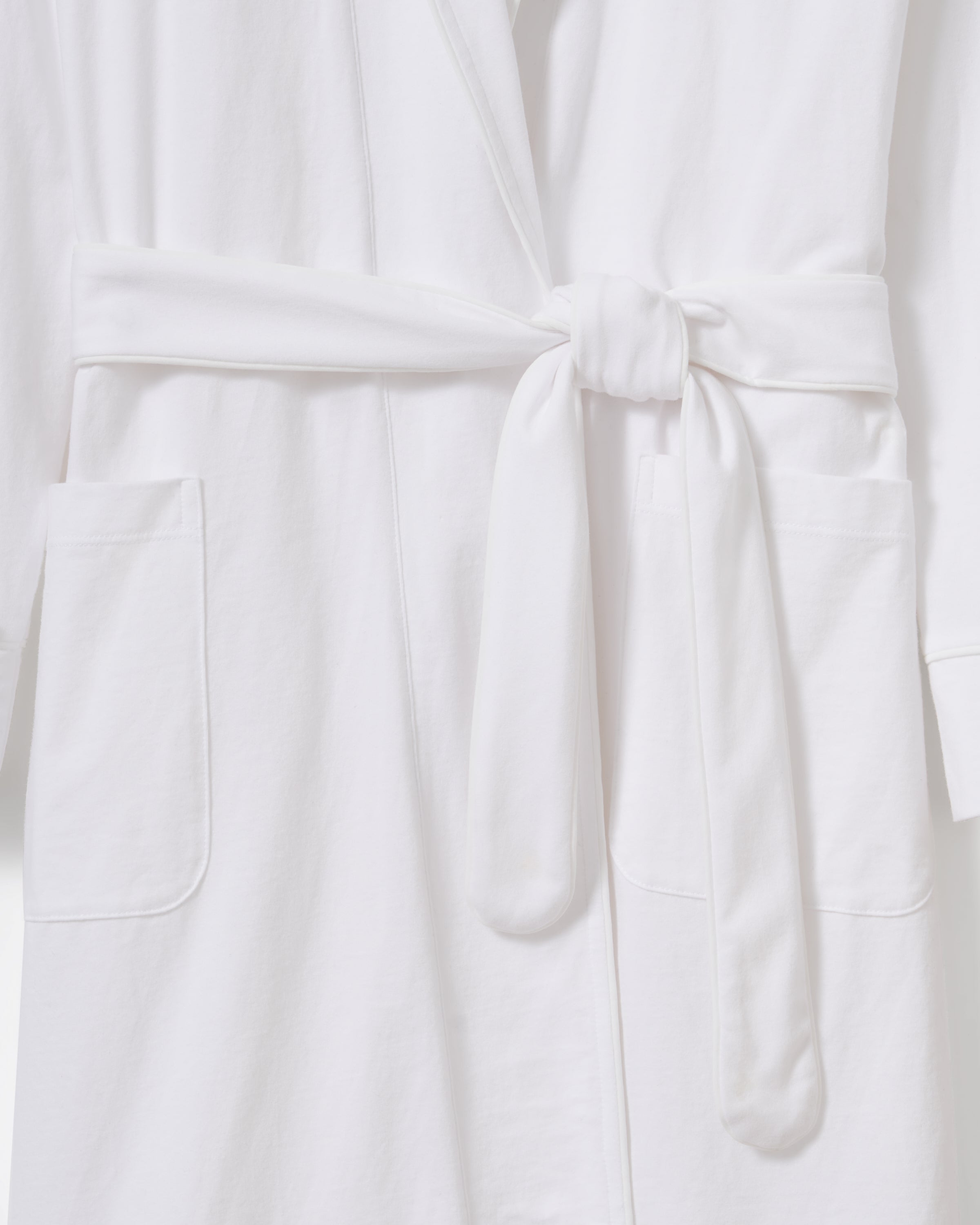 A person with curly hair is wearing a Petite Plume Womens Pima Robe in White, crafted from luxurious Peruvian Pima cotton. With hands in pockets, they stand against a plain light background, looking forward with a calm expression.