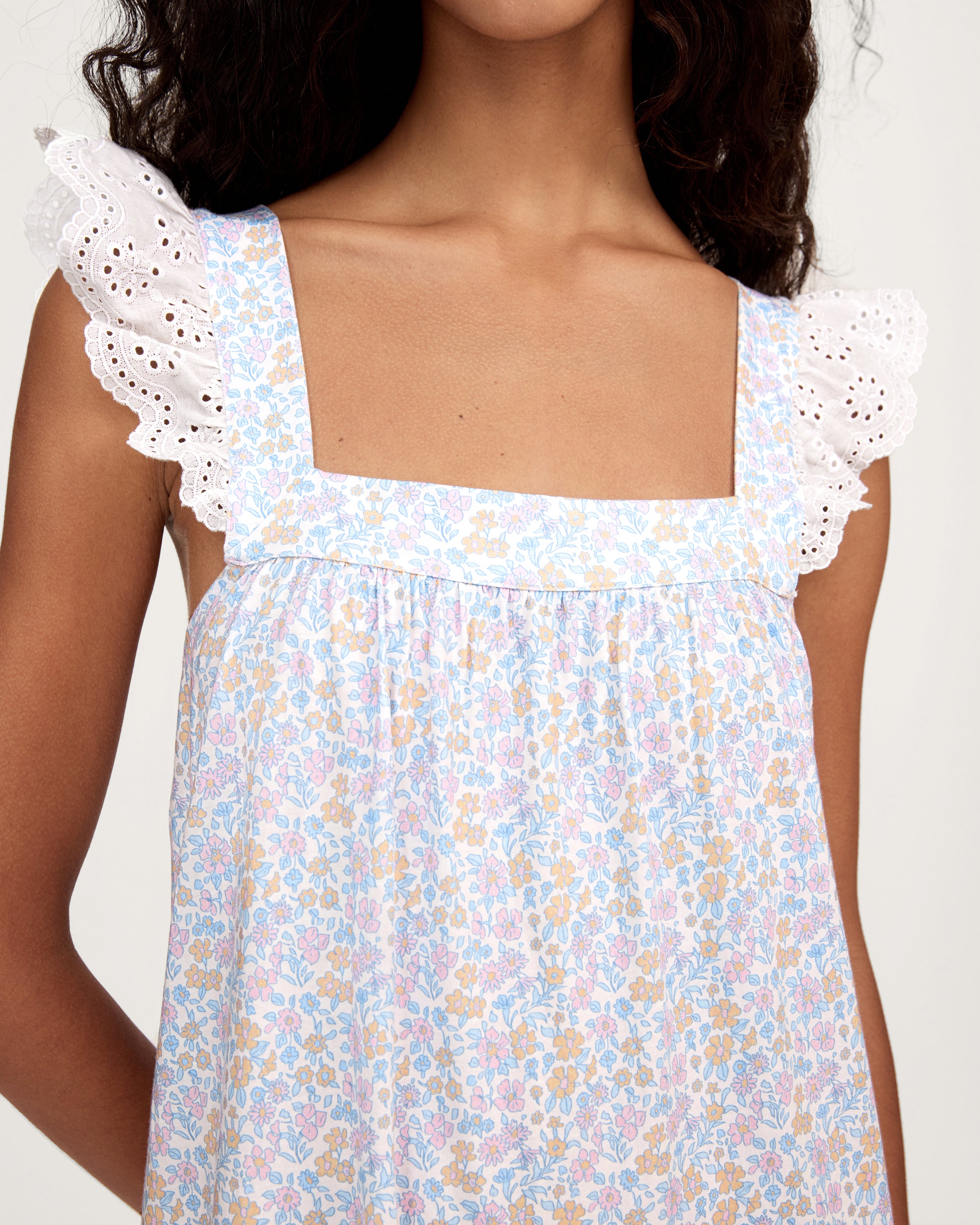 A woman with long curly hair stands barefoot on a plain light background, wearing the Women's Lightweight Pima Clara Nightgown in Fleur Blanche—an ankle-length, sleeveless gown with dainty floral print and lace-trimmed straps.