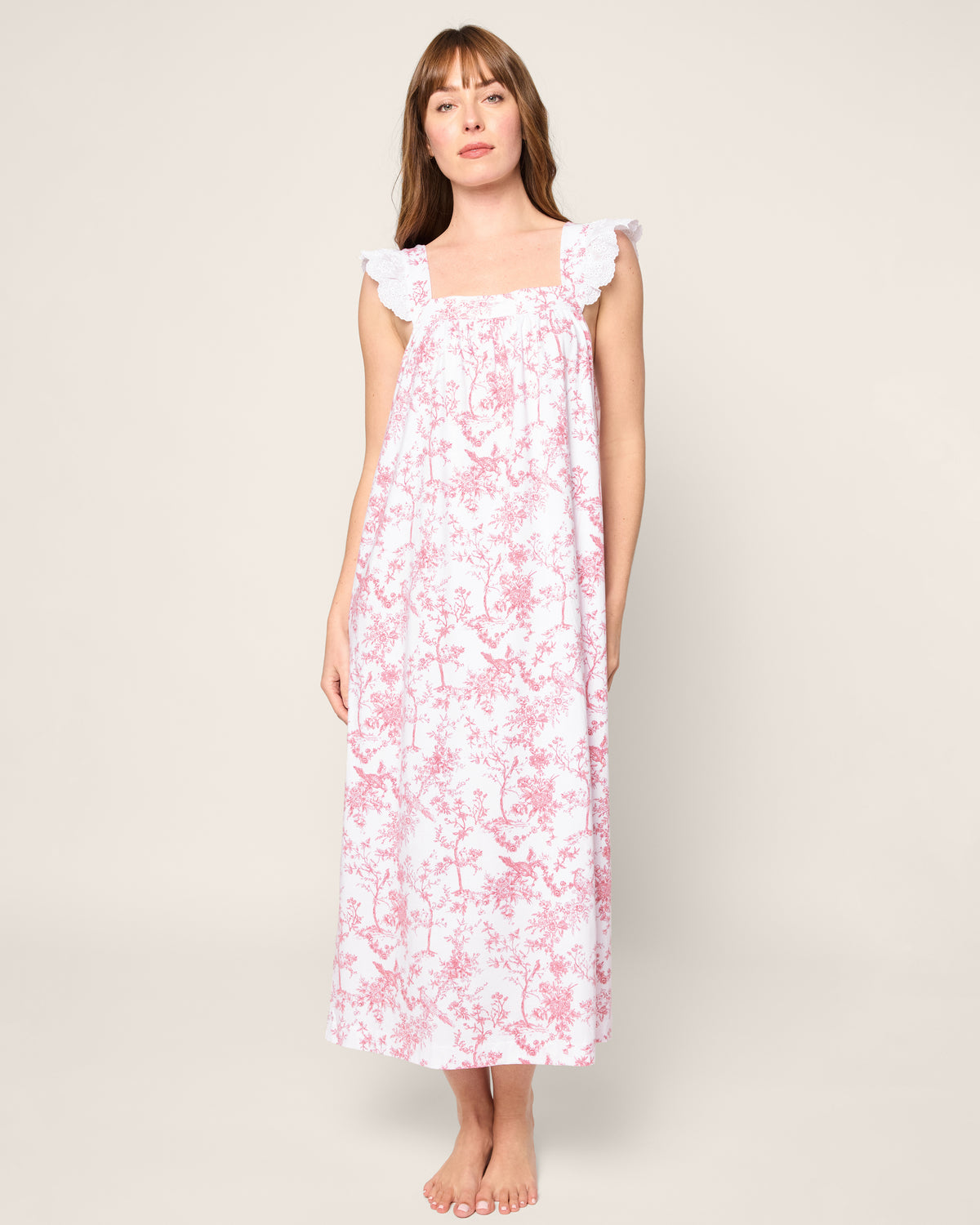 A person stands barefoot in a sleeveless Women’s Pima Clara Nightgown in Pink Timeless Toile by Petite Plume. The dress, with lace strap details and floral pattern, resembles luxurious pajamas perfect for bedtime. The background is a plain light color.
