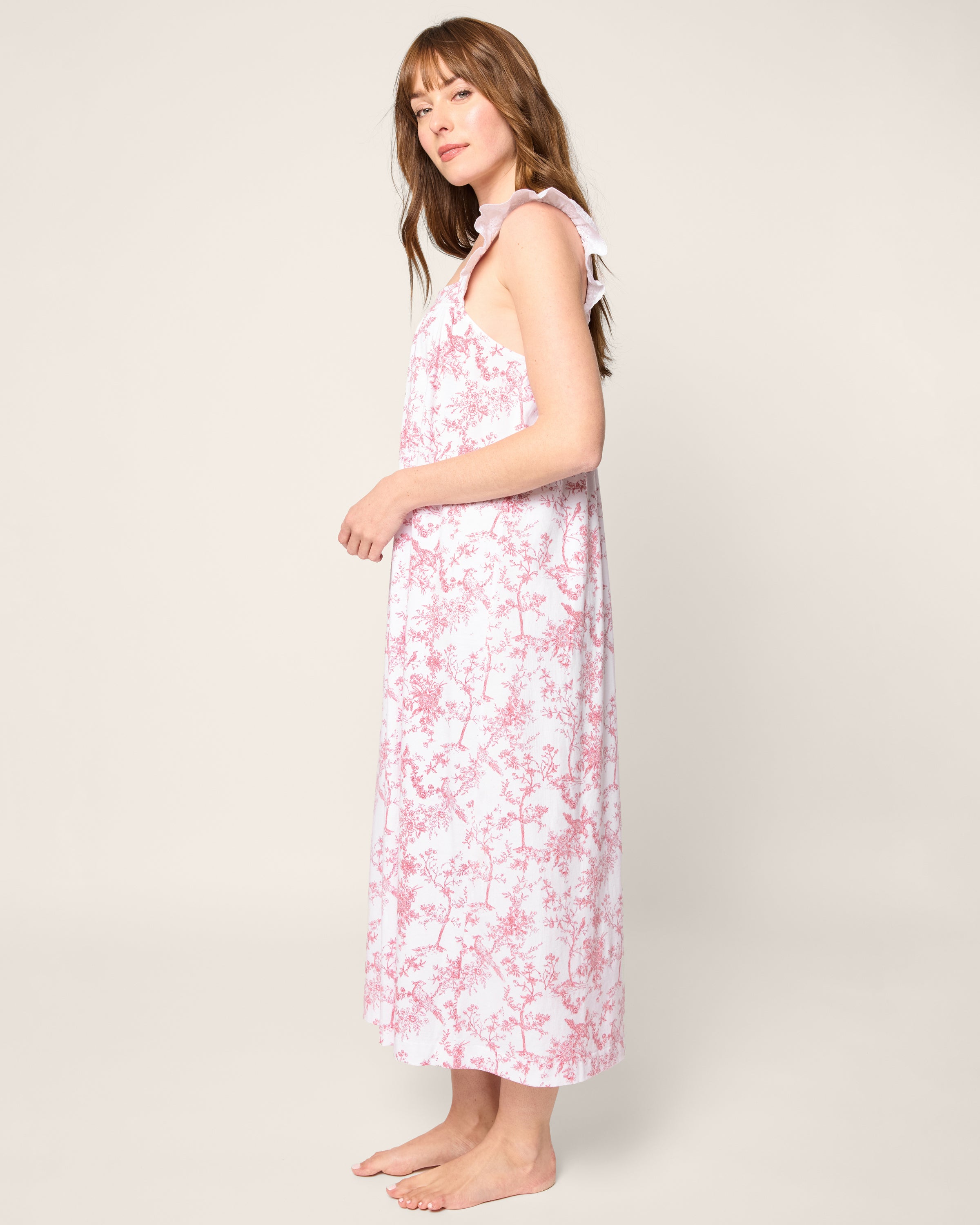 A person with long hair stands sideways barefoot, exuding serenity in Petite Plumes Womens Pima Clara Nightgown in Pink Timeless Toile, featuring a white and pink floral design with tied shoulder straps, resembling luxurious pajamas.