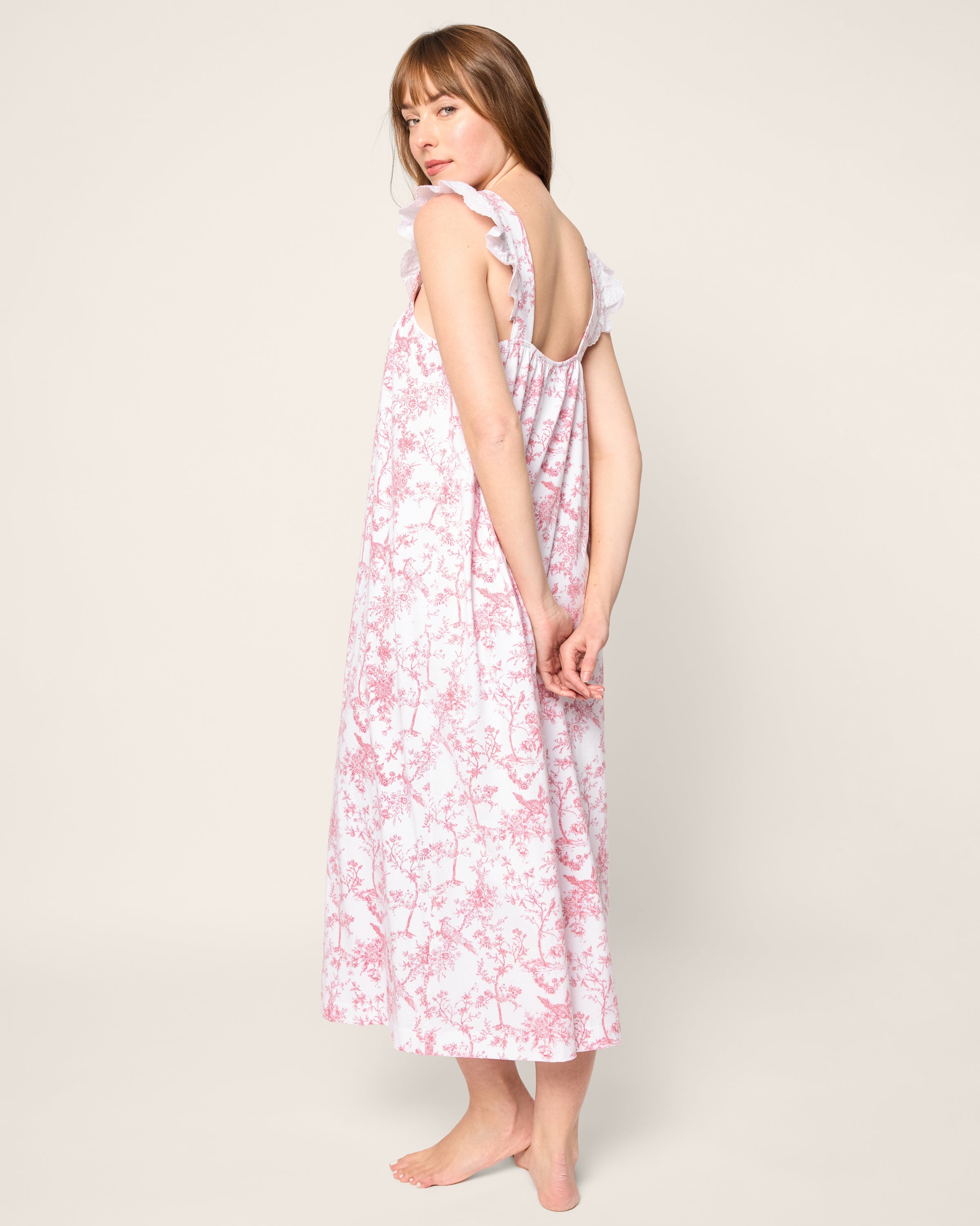 Barefoot on a light background, a person models the Womens Pima Clara Nightgown in Pink Timeless Toile by Petite Plume. They face slightly away, one arm down and the other touching their hair. The nightgowns ruffled straps enhance its elegant charm, resembling luxurious pajamas.