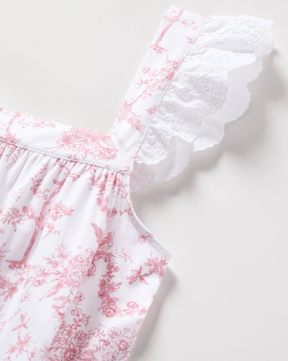 The Womens Pima Clara Nightgown in Pink Timeless Toile by Petite Plume showcases a pink floral pattern with a ruffled shoulder strap. The nightgowns fabric features delicate pink designs, reminiscent of luxurious pajamas, with elegant white lace detailing on the strap.