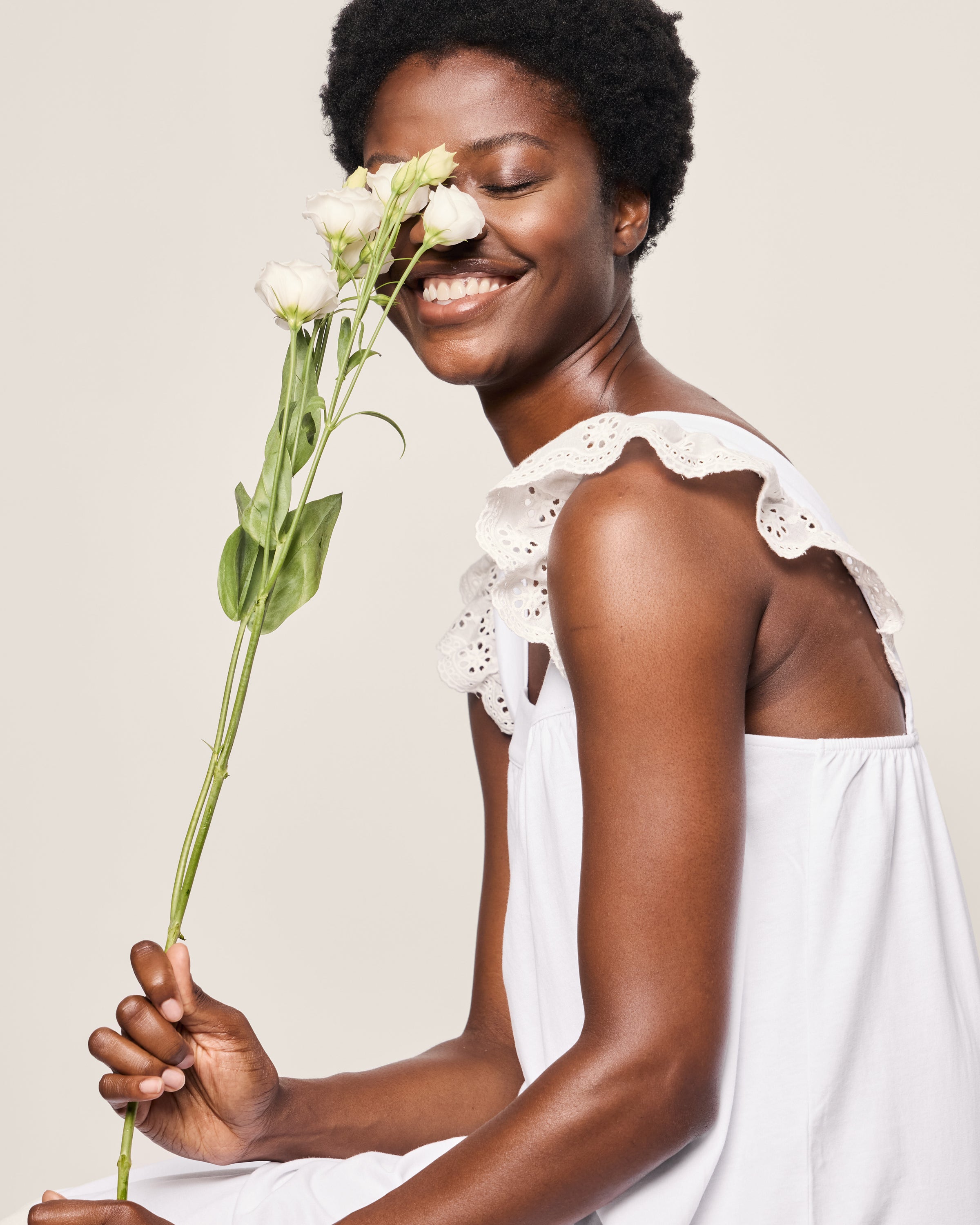 A person smiles while holding white flowers near their face, wearing Petite Plumes Womens Pima Clara Nightgown in White with lace details, against a neutral light background, evoking the serenity of a bedtime ritual.