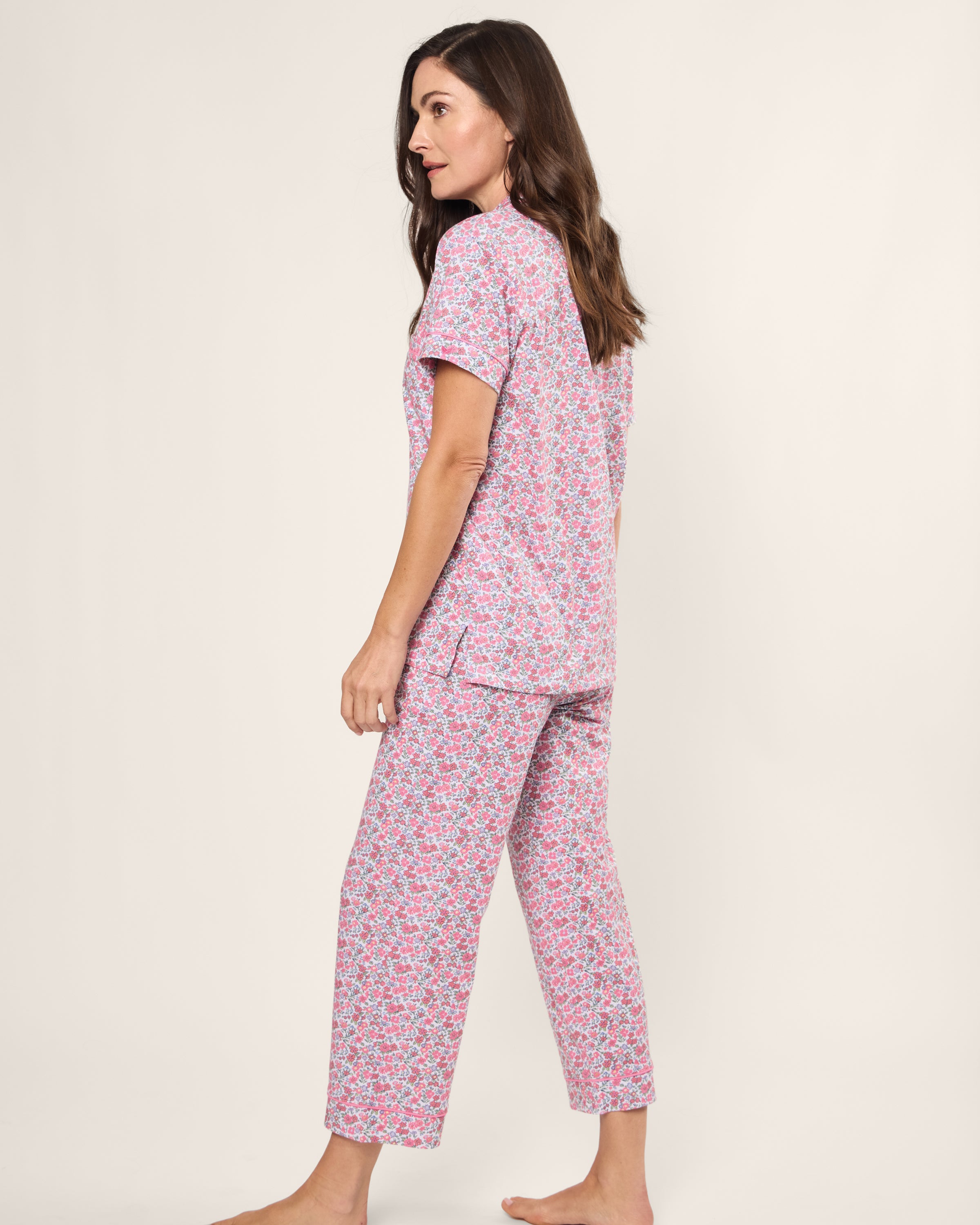 A woman in the Womens Pima Crop Set in Fleurs de Rose by Petite Plume stands barefoot, sporting pink floral pajamas with a side view against a plain backdrop.