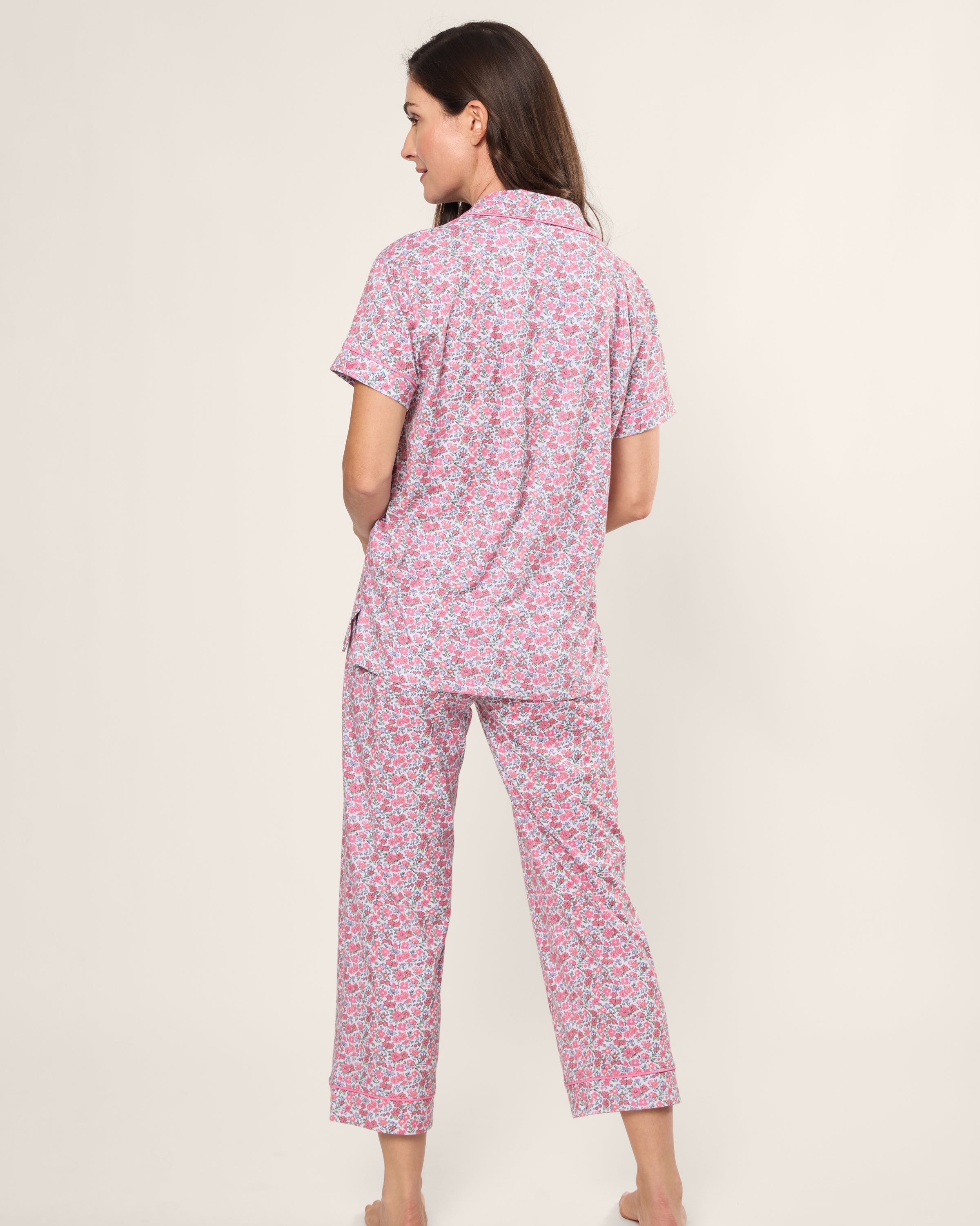A woman with long brown hair stands barefoot, facing away, wearing a Womens Pima Crop Set in Fleurs de Rose by Petite Plume. The vintage-style pink floral ensemble includes short sleeves and long pants, set against a plain light background.