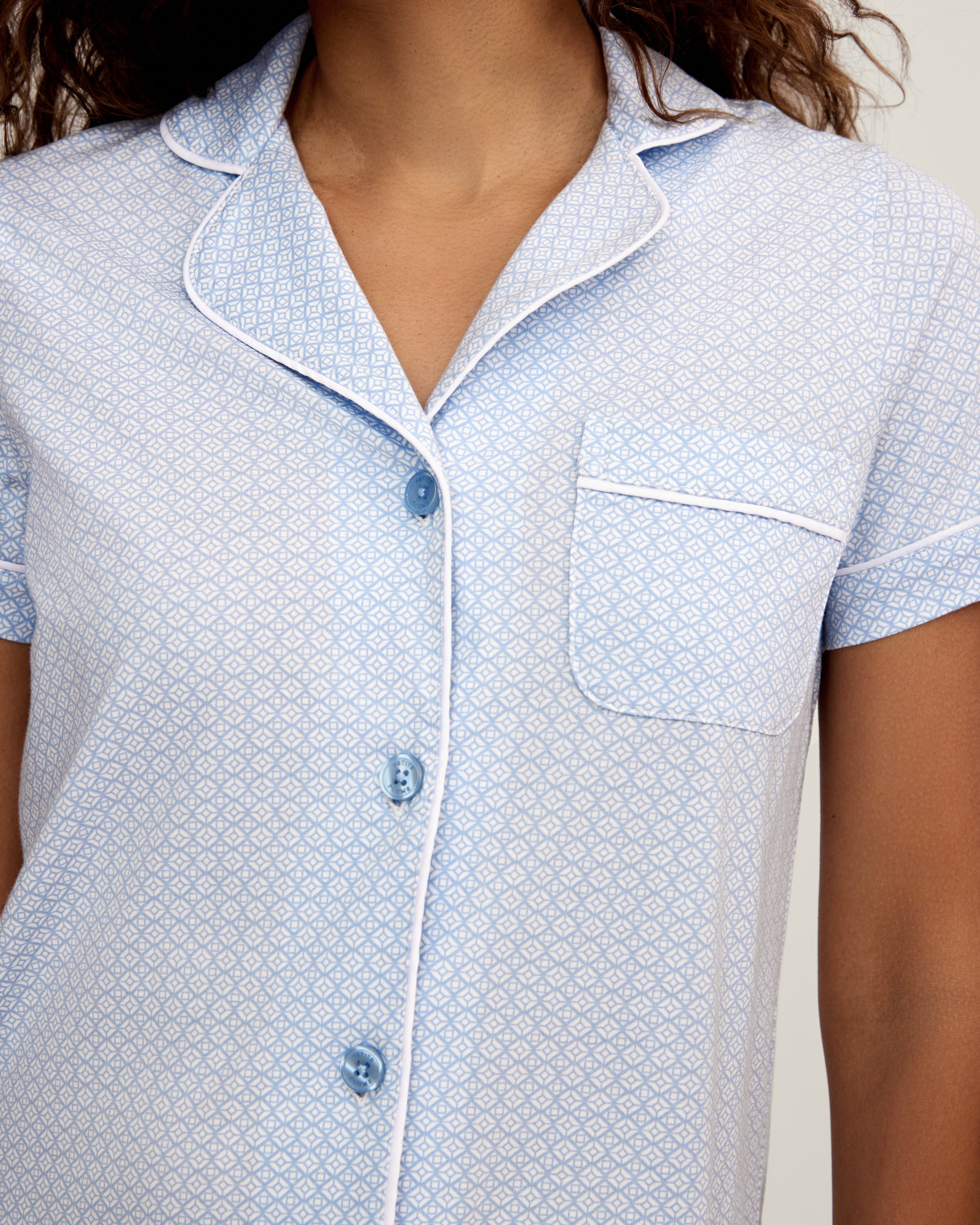 A woman stands barefoot in the Women's Pima Crop Set in Periwinkle Geo—light blue, short-sleeve, button-up pajamas with a collar, chest pocket, and matching loose-fitting pants—against a plain light background.