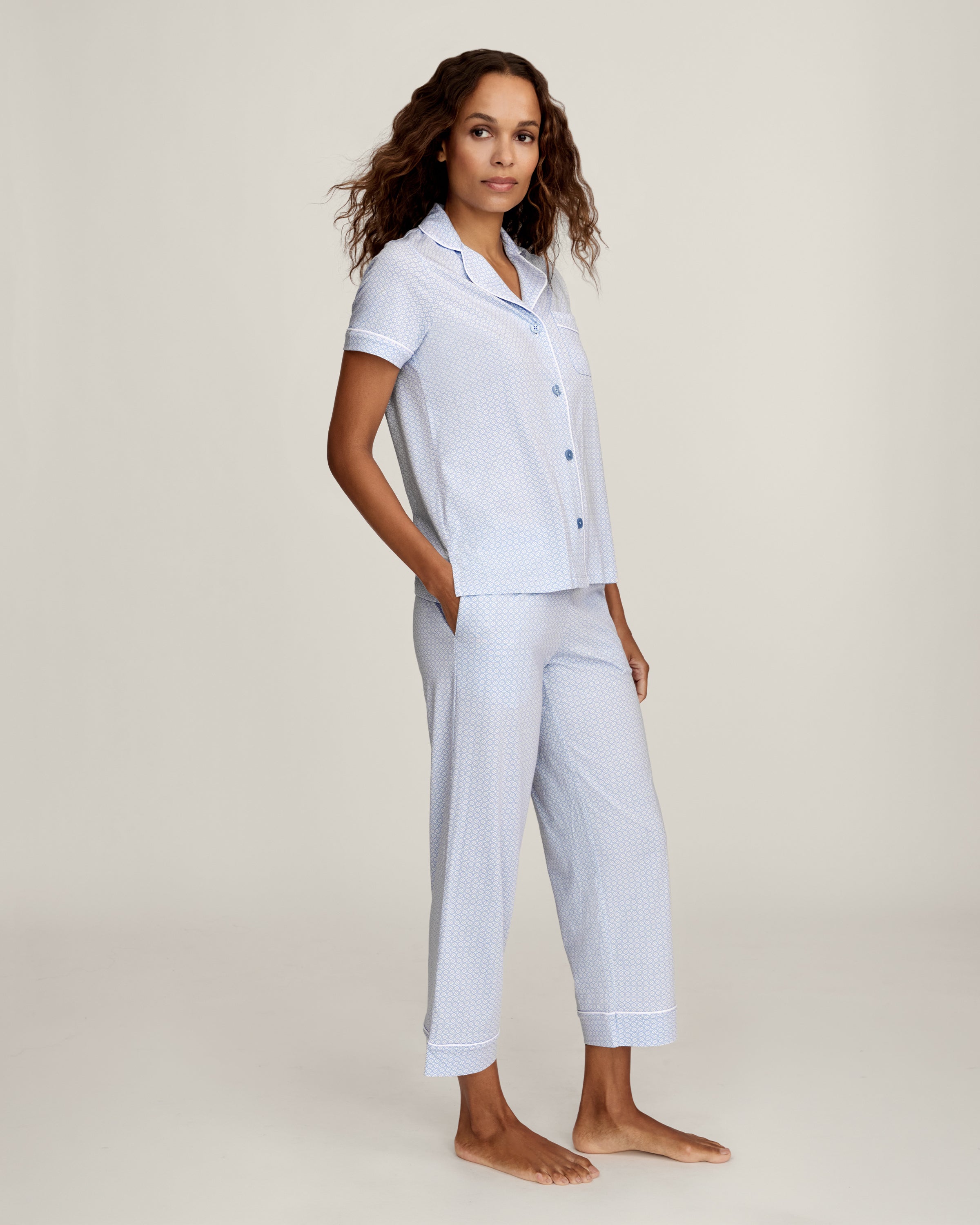 A woman stands barefoot in the Women's Pima Crop Set in Periwinkle Geo—light blue, short-sleeve, button-up pajamas with a collar, chest pocket, and matching loose-fitting pants—against a plain light background.