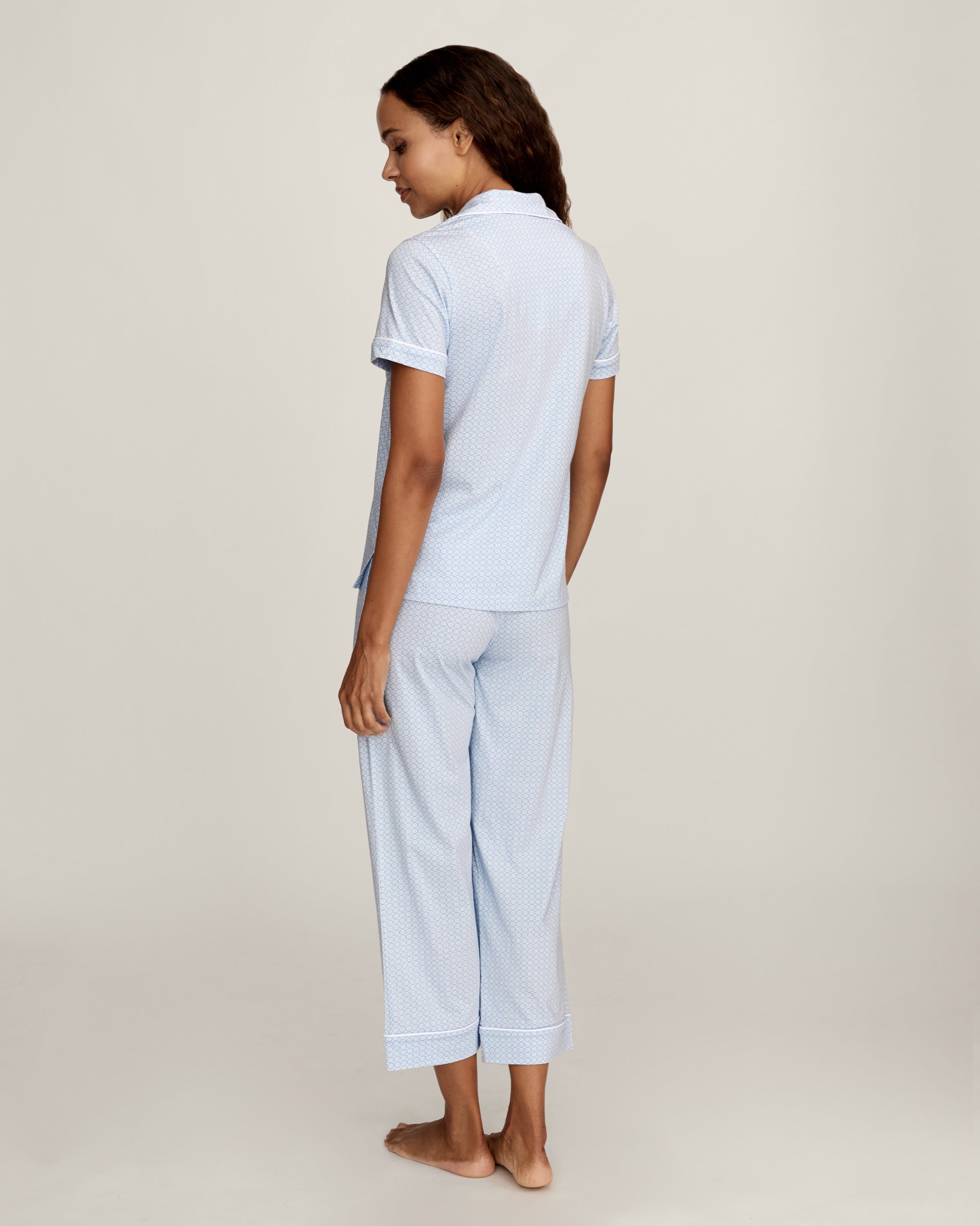 A woman stands barefoot in the Women's Pima Crop Set in Periwinkle Geo—light blue, short-sleeve, button-up pajamas with a collar, chest pocket, and matching loose-fitting pants—against a plain light background.