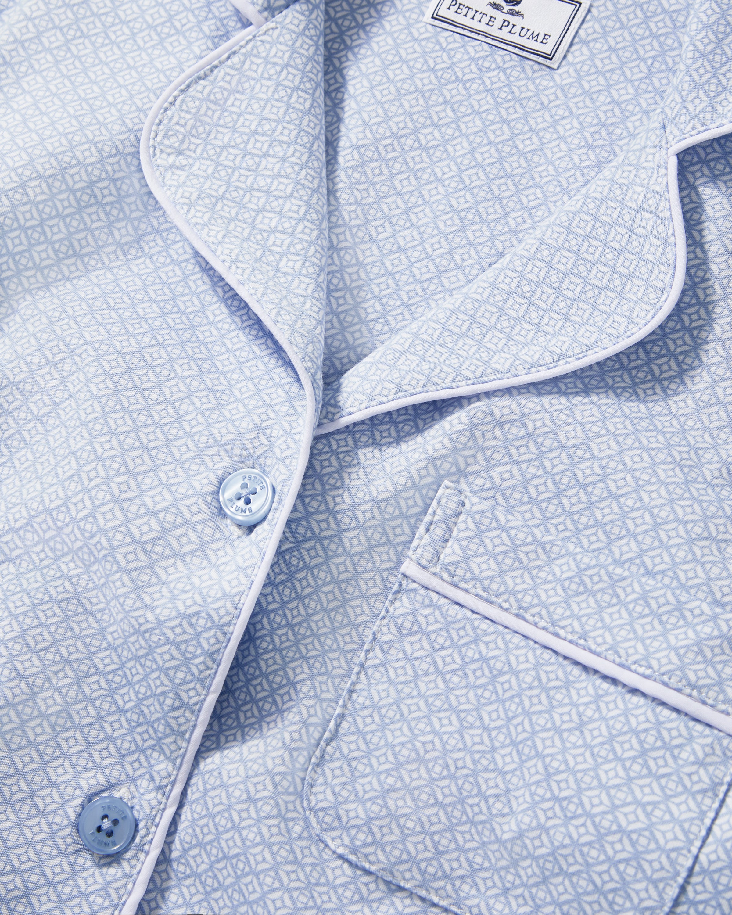 A woman stands barefoot in the Women's Pima Crop Set in Periwinkle Geo—light blue, short-sleeve, button-up pajamas with a collar, chest pocket, and matching loose-fitting pants—against a plain light background.