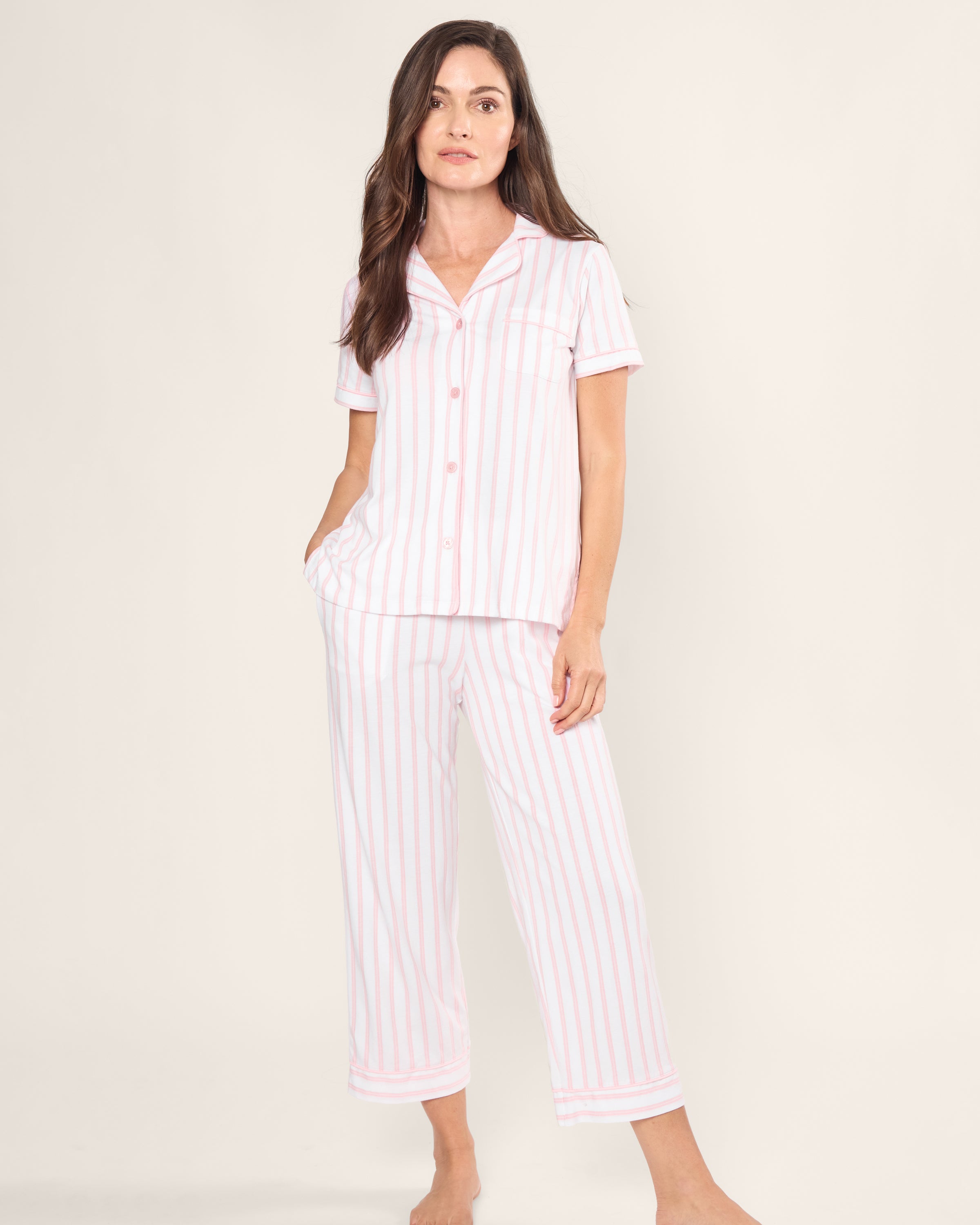 A person stands barefoot in the Womens Pima Crop Set by Petite Plume, featuring a vintage-inspired short-sleeved, button-up top with a collar and cropped pants. Made from soft Peruvian Pima cotton, it displays pink and white stripes against a plain white background.
