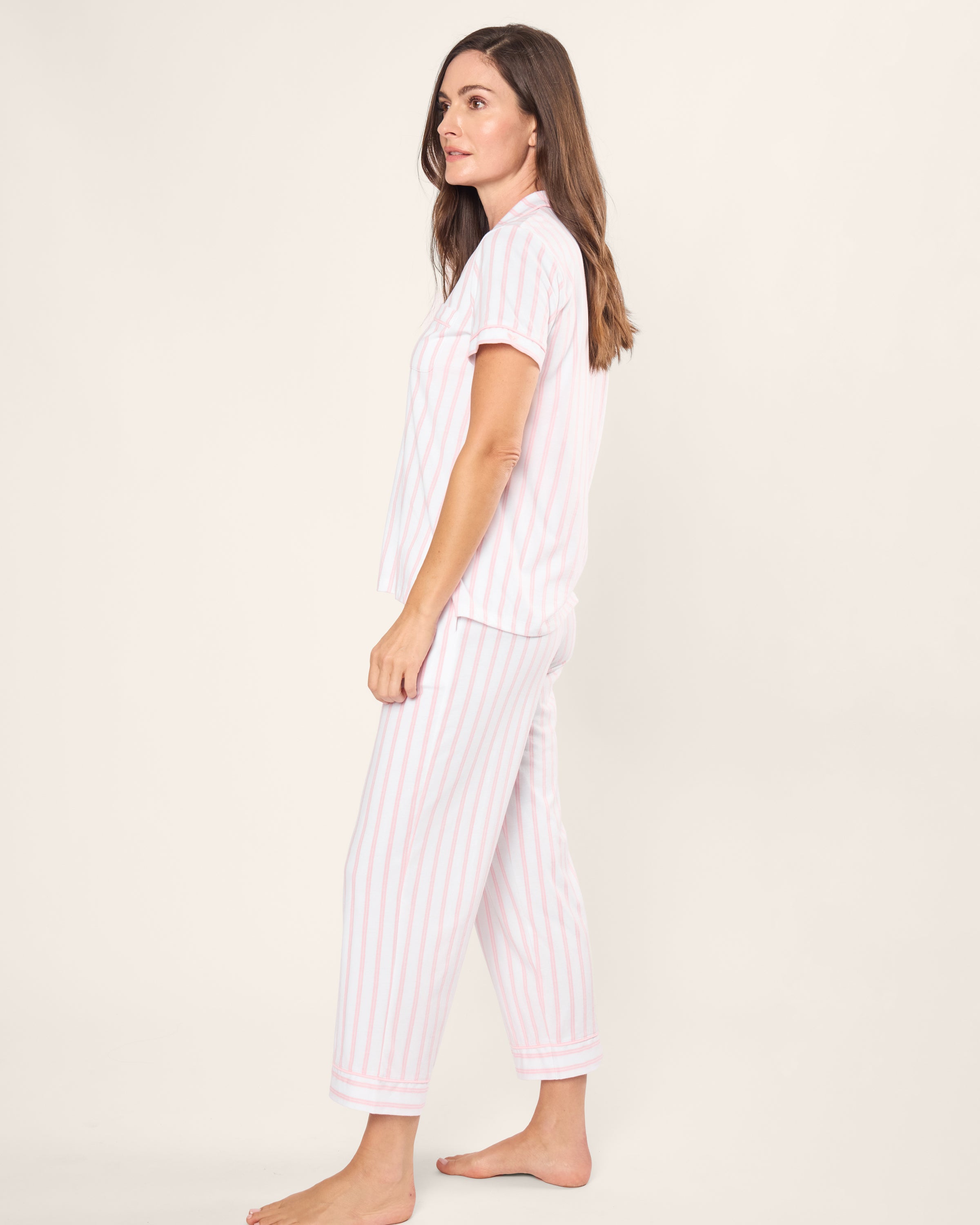 A long-haired brunette stands barefoot in the Womens Pima Crop Set by Petite Plume, featuring pink and white stripes. She faces sideways against a plain white background, looking slightly upward.