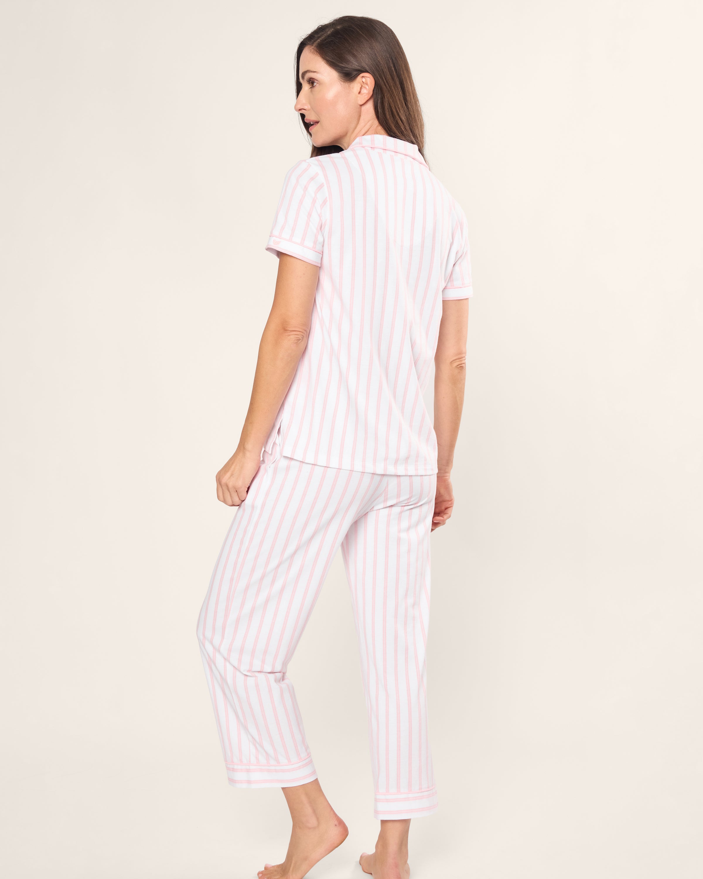 A woman stands barefoot in Petite Plumes Womens Pima Crop Set in Pink and White Stripe. Crafted from soft Pima cotton, she wears a short-sleeve button-up shirt and cropped pants against a plain off-white background.