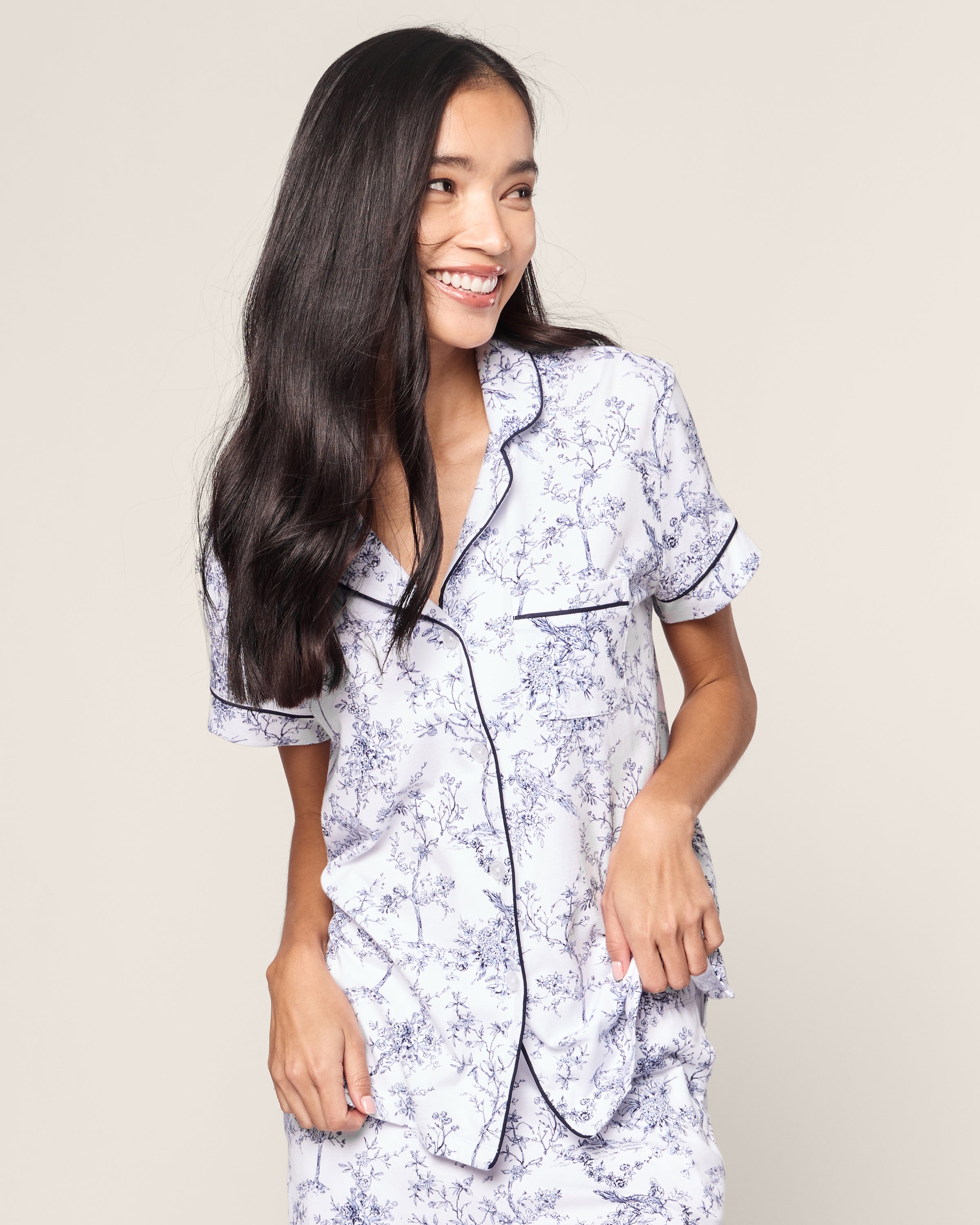 A woman with long dark hair smiles gracefully against a plain backdrop, wearing the Womens Pima Crop Set in Timeless Toile by Petite Plume. The pajama set features an elegant floral pattern and dark piping, with short sleeves and a classic collar for a timeless feel.