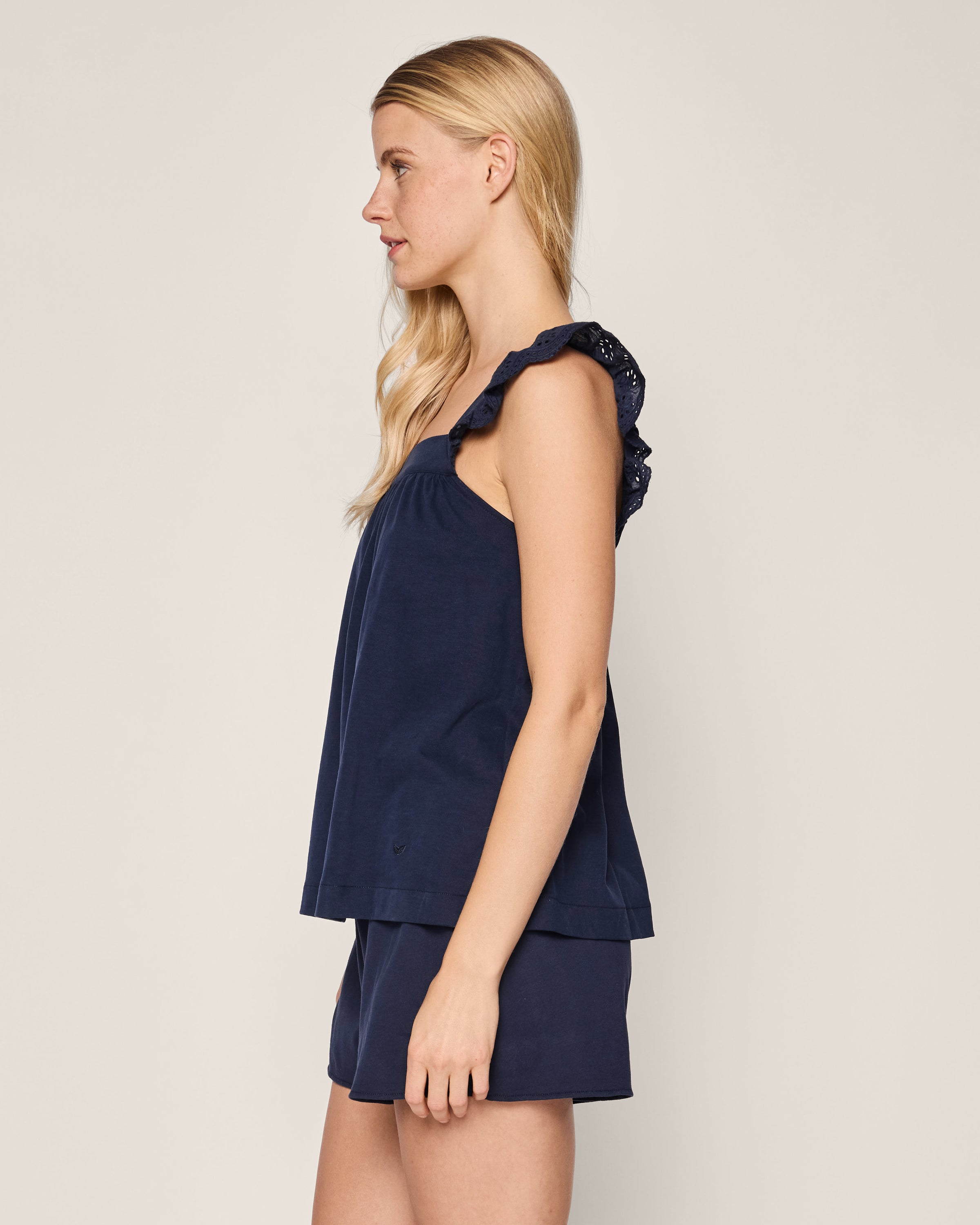A woman with long blonde hair is in profile, wearing Petite Plumes Womens Pima Clara Short Set in navy, featuring a soft sleeveless top with ruffled straps and matching shorts, set against a simple light background, capturing the essence of an elegant luxury bedtime ritual.