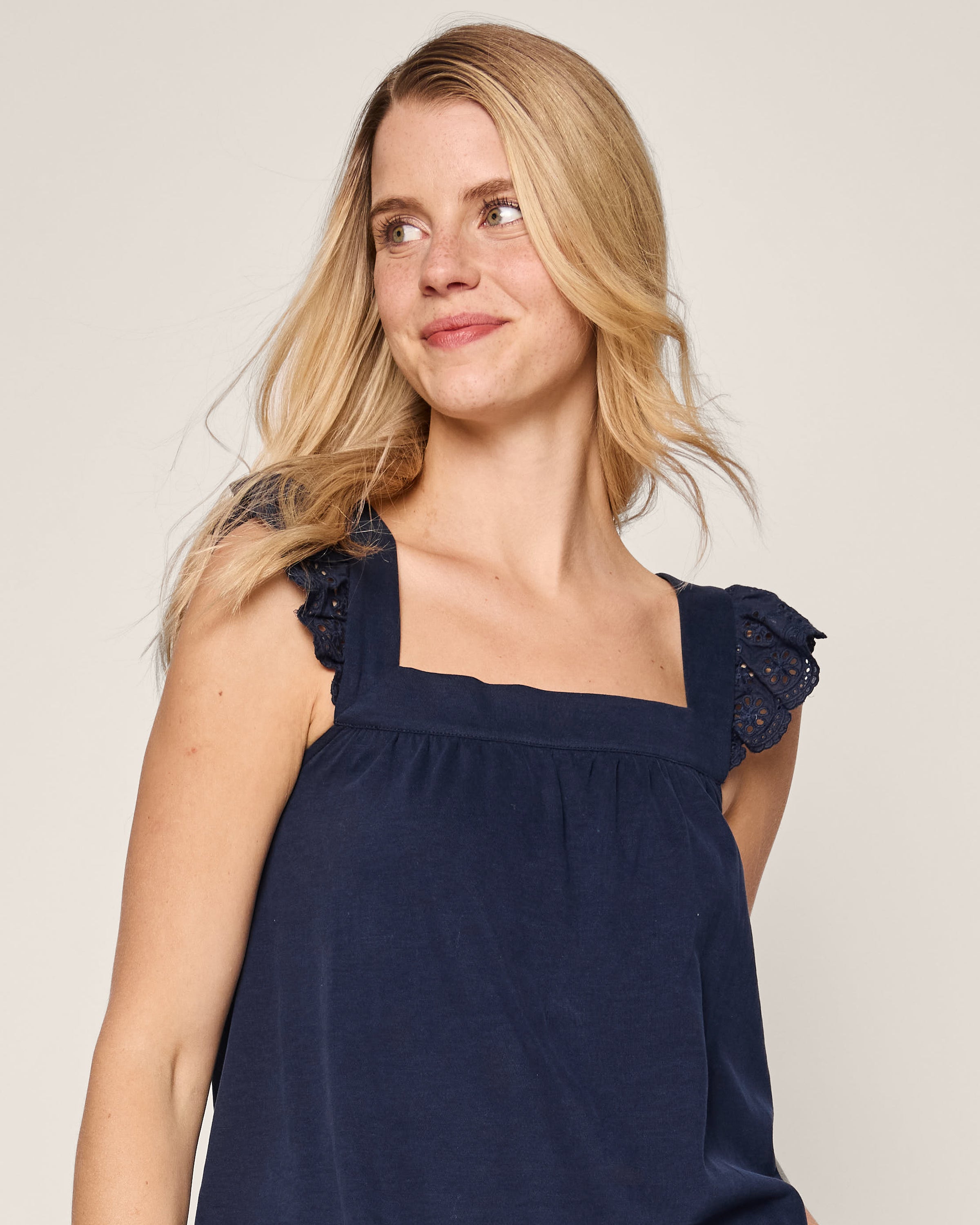 A woman with long blonde hair smiles, glancing sideways. Shes wearing a navy sleeveless top with lace shoulder details, similar to Petite Plumes Womens Pima Clara Short Set in Navy. The background is plain and light-colored.
